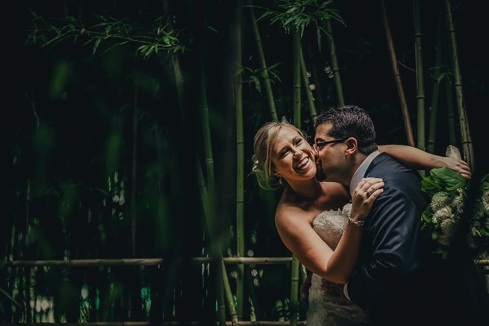 A newlywed couple sharing a kiss and laughing in a lush, green bamboo forest at night.