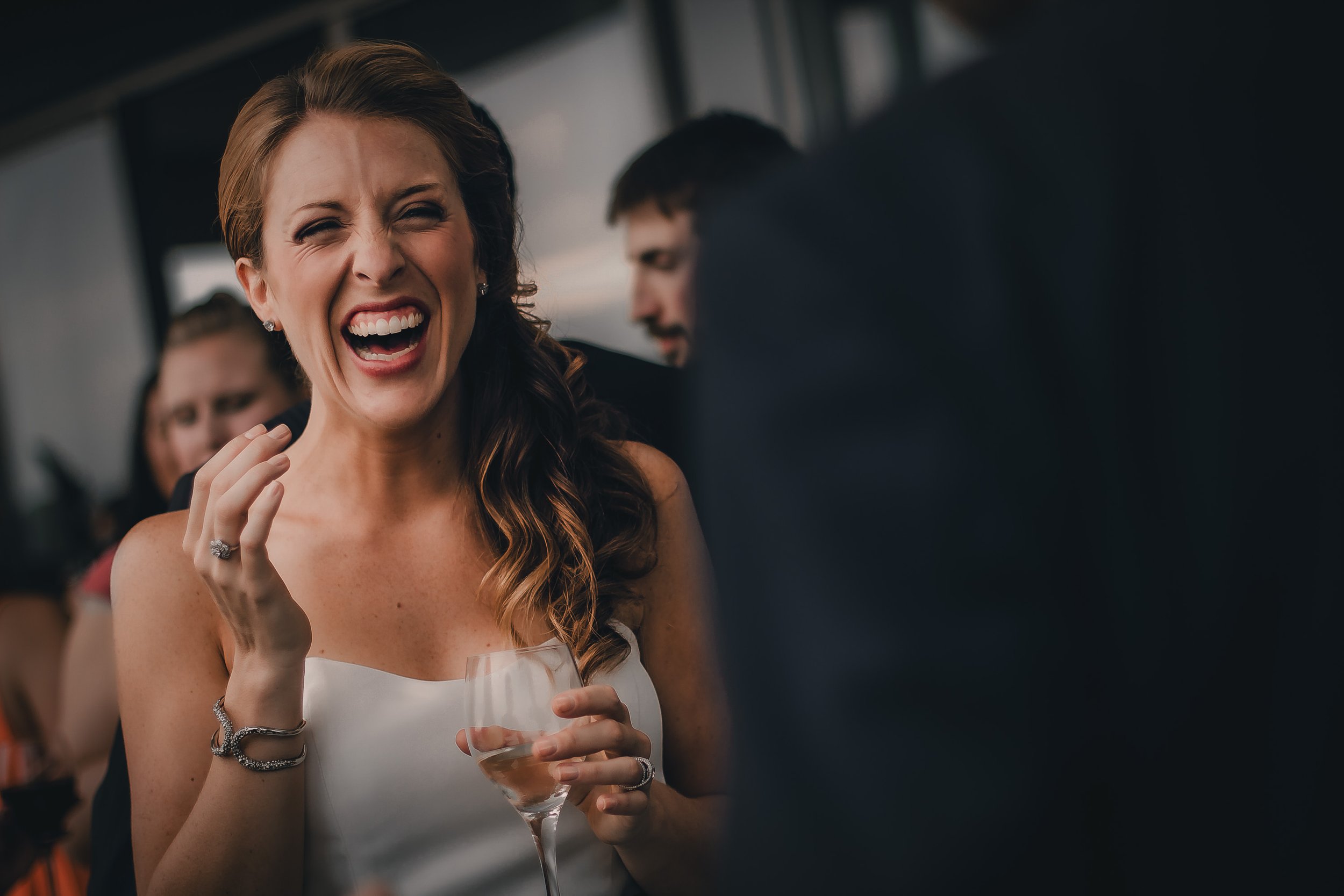 A woman in a white dress laughing and holding a glass of wine at a social event, with other people in the background.