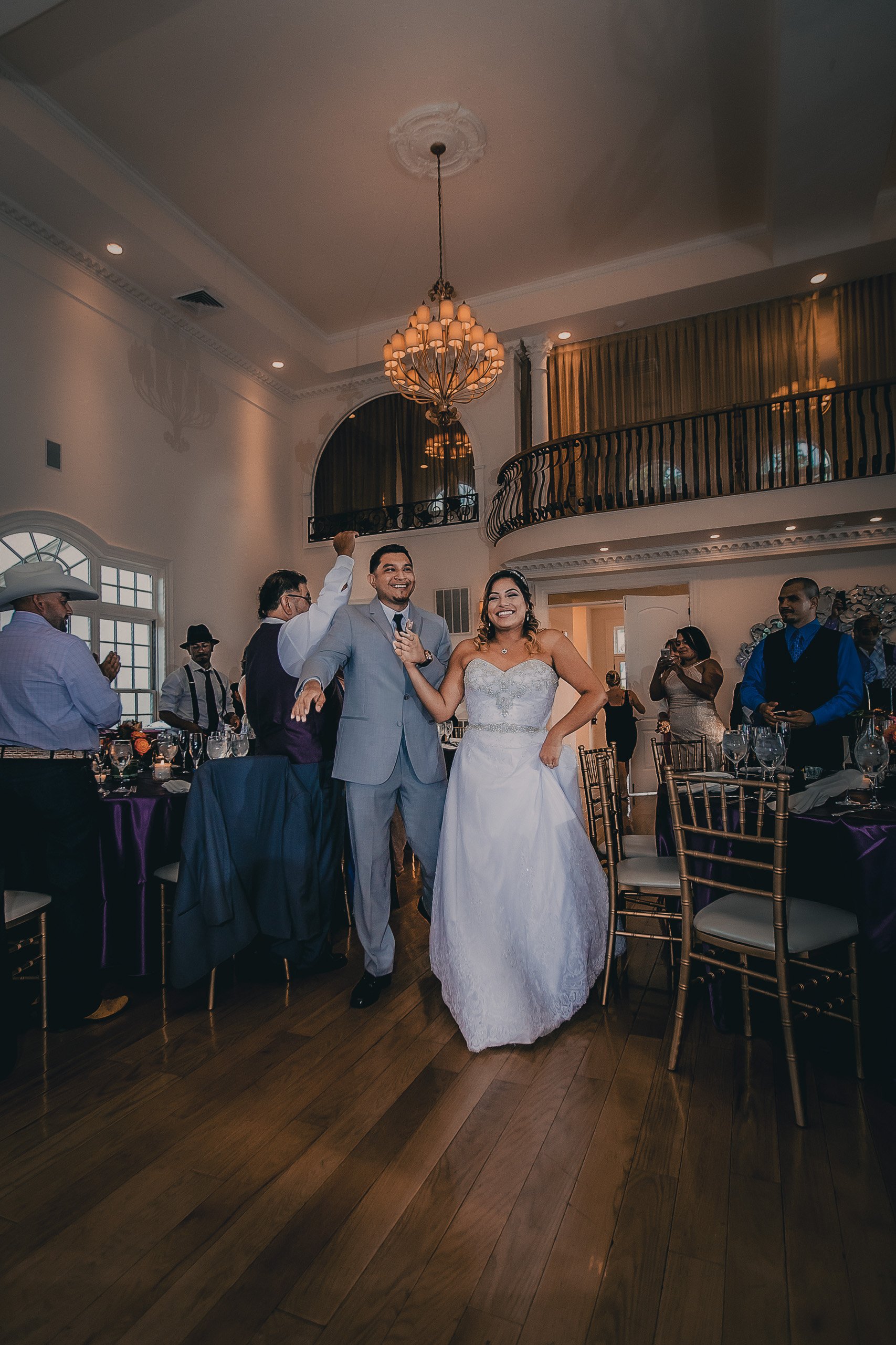 Bride and groom smiling and celebrating during wedding reception in a decorated elegant banquet hall with guests seated around tables.