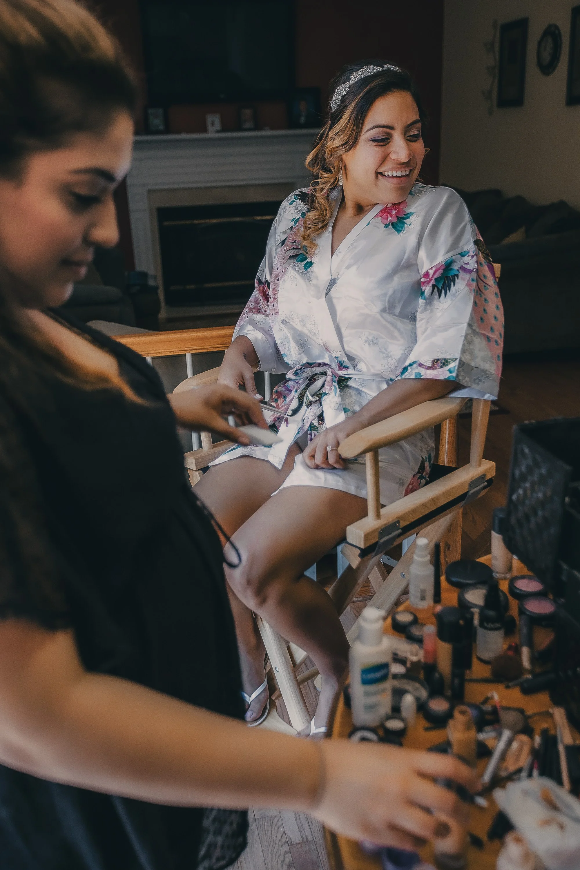 A woman in bridal robes having her legs or feet painted or having a makeup treatment, with makeup products and tools on a table nearby.