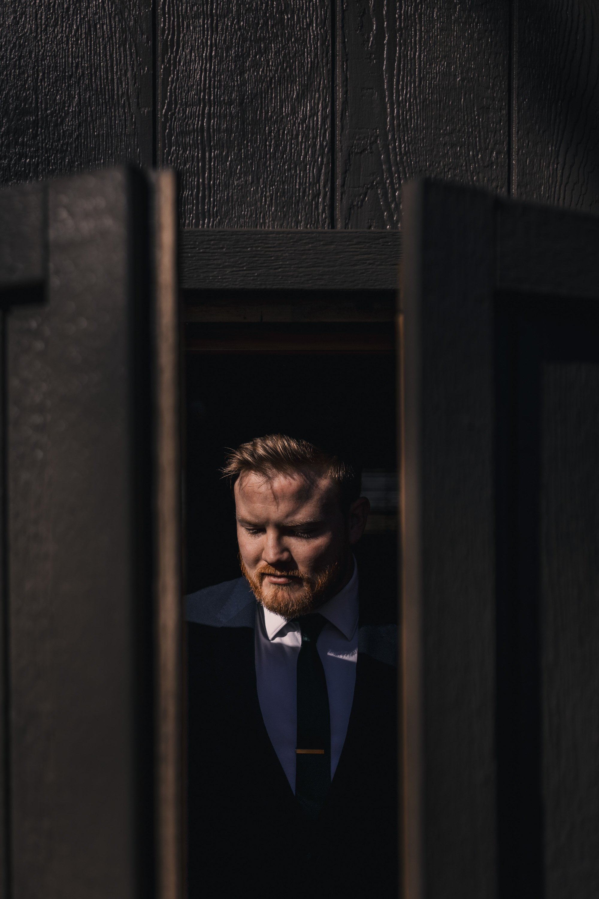 A man with a beard and brown hair looking down, dressed in a suit and tie, seen through an opening in a dark wooden structure.