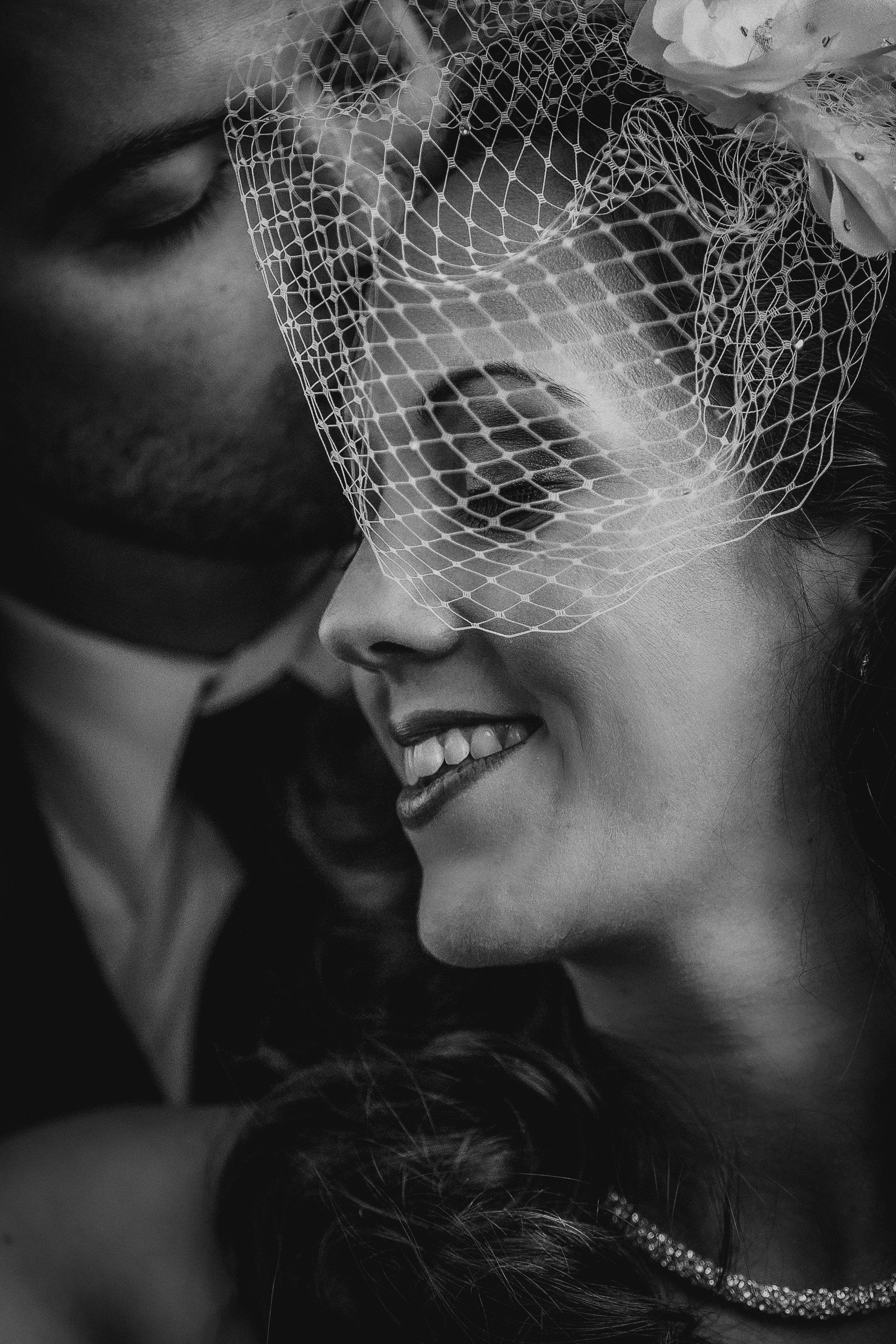 A close-up black and white photograph of a couple smiling, with the woman wearing a lace veil with floral details over her face, and the man's face close to hers, both smiling.