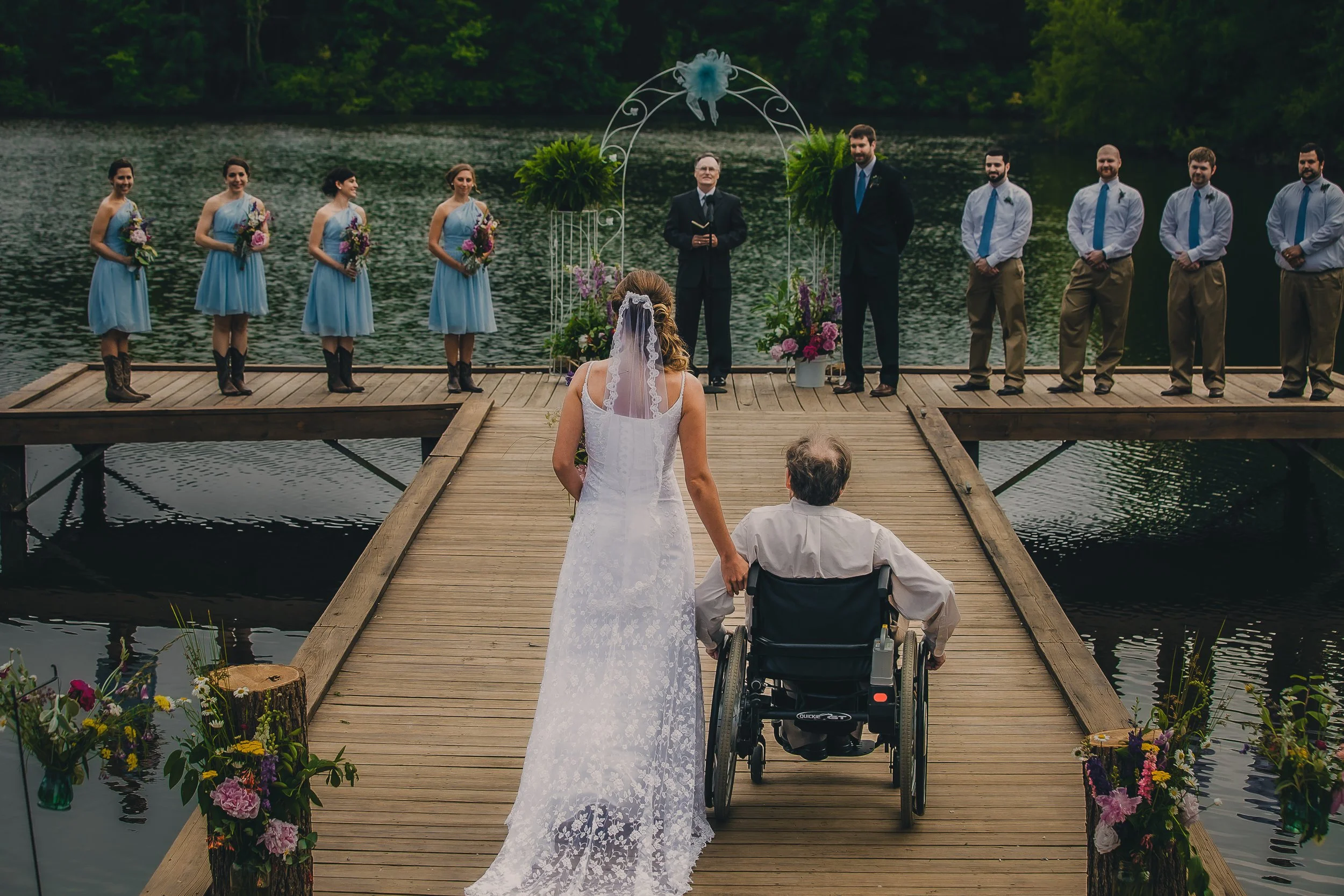A bride in a white lace wedding dress walking with a man in a wheelchair toward an outdoor wedding ceremony on a wooden dock by a lake, with bridesmaids in blue dresses and groomsmen in white shirts with blue ties.
