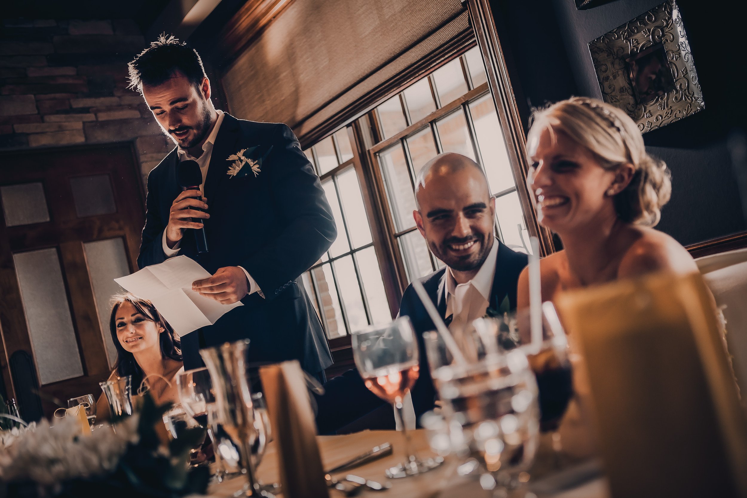 A man giving a speech at a wedding reception with guests listening, including a woman smiling and a man and woman seated at the table.
