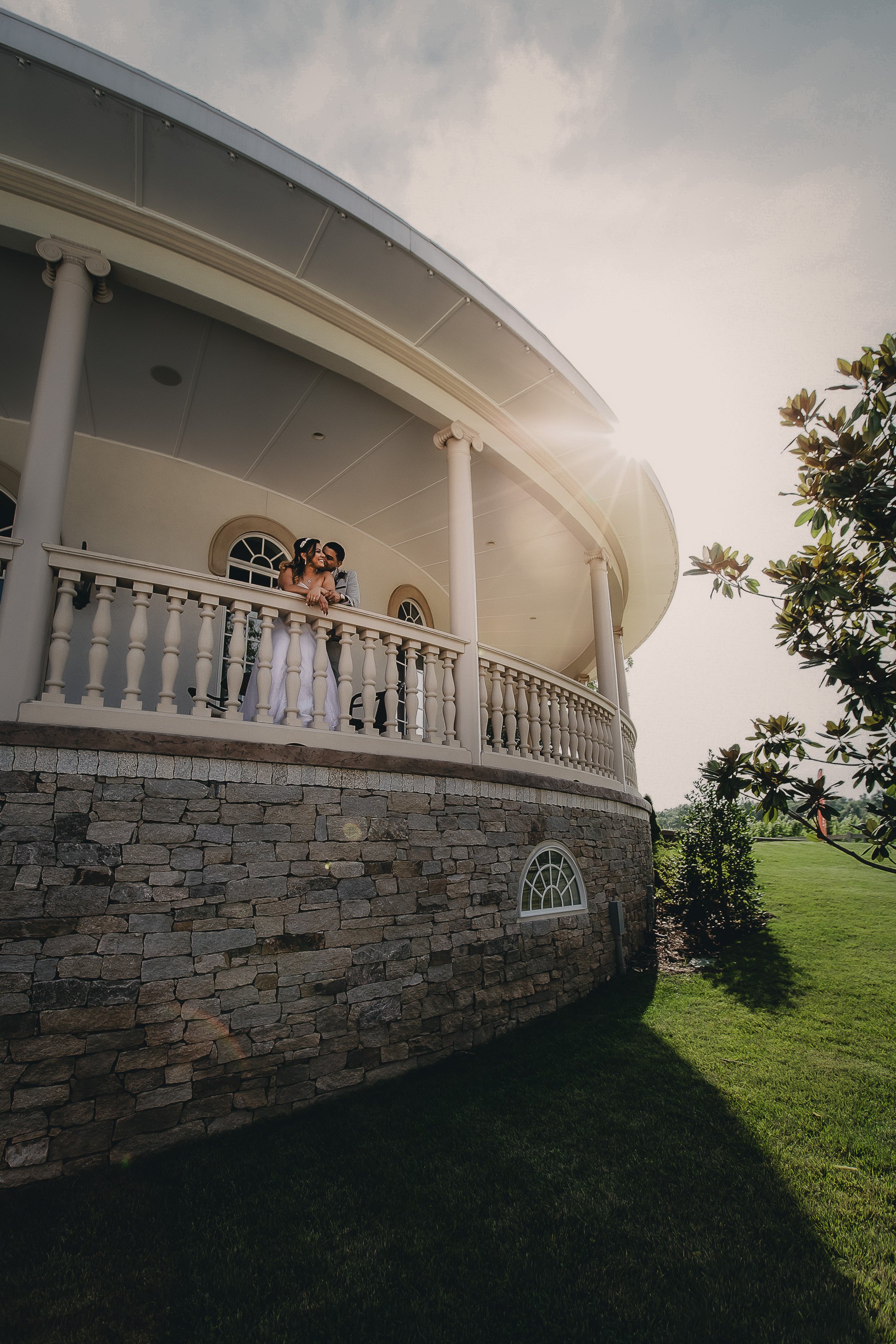 A couple is standing on a large, curved balcony of a white house with stone foundation, surrounded by green grass and trees, with sunlight shining from behind.