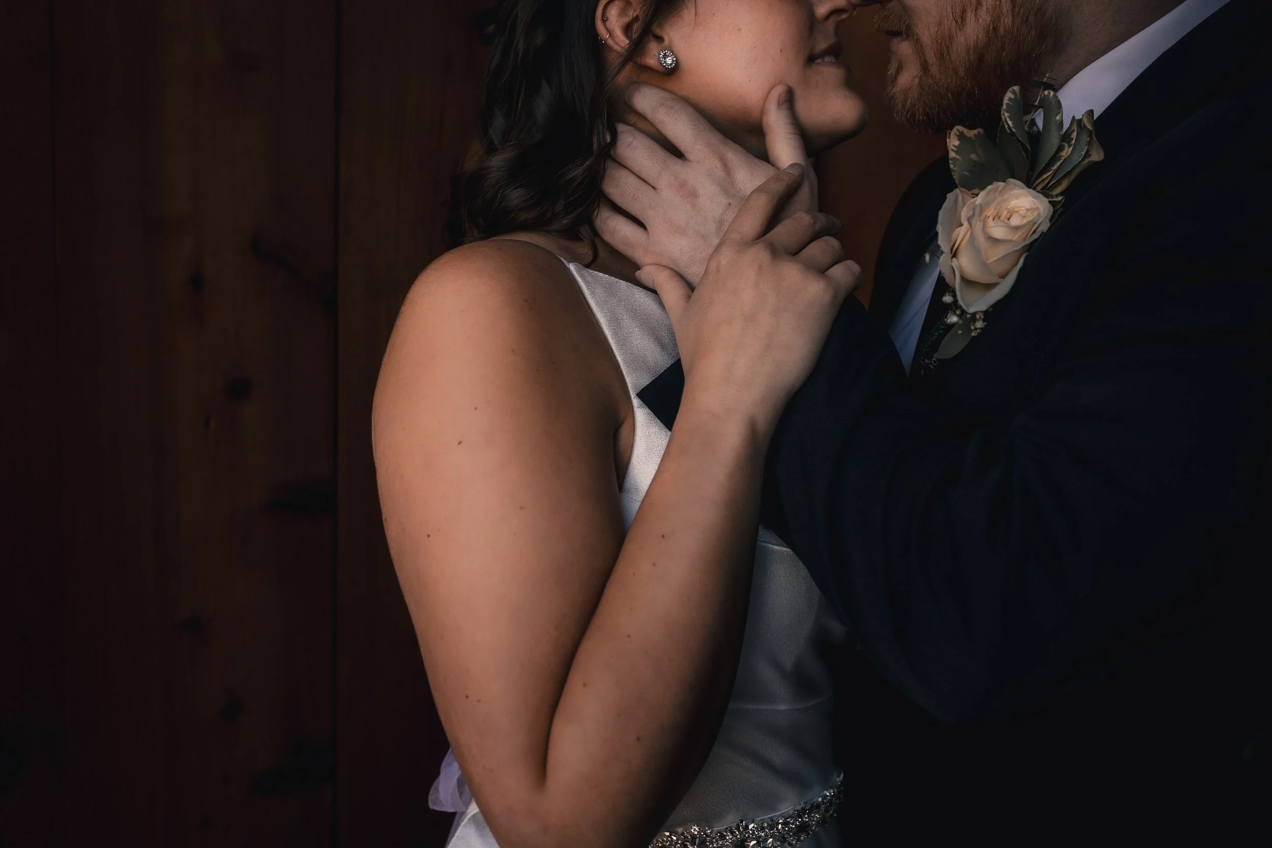 A close-up of a couple embracing closely, with the woman's face near the man's face, and the woman's hand gently touching the man's neck. The woman is wearing earrings and a satin dress, and the man is dressed in a suit with a boutonniere.