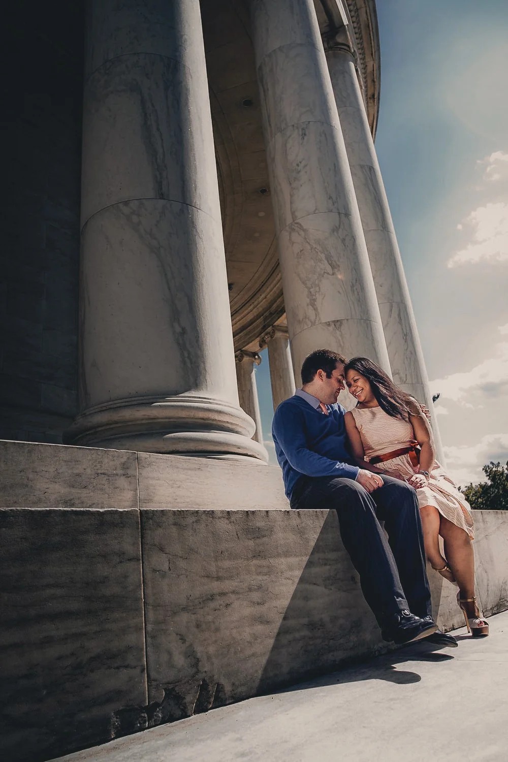 Couple sitting closely together on a marble ledge in front of large white marble columns of a classical building, smiling and touching foreheads, under a partly cloudy sky.