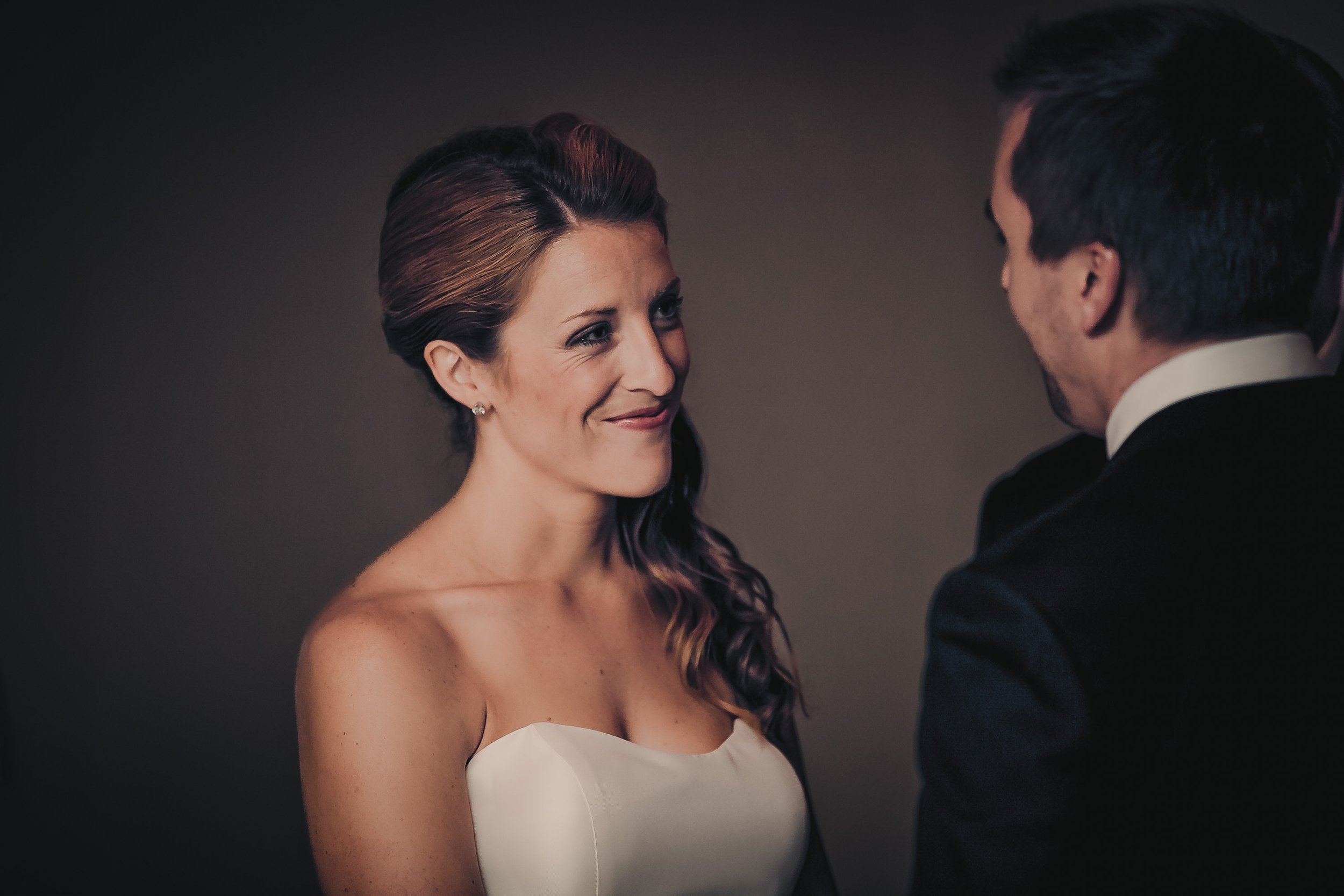 A woman in a strapless white dress smiling at a man in a black suit during a wedding ceremony.