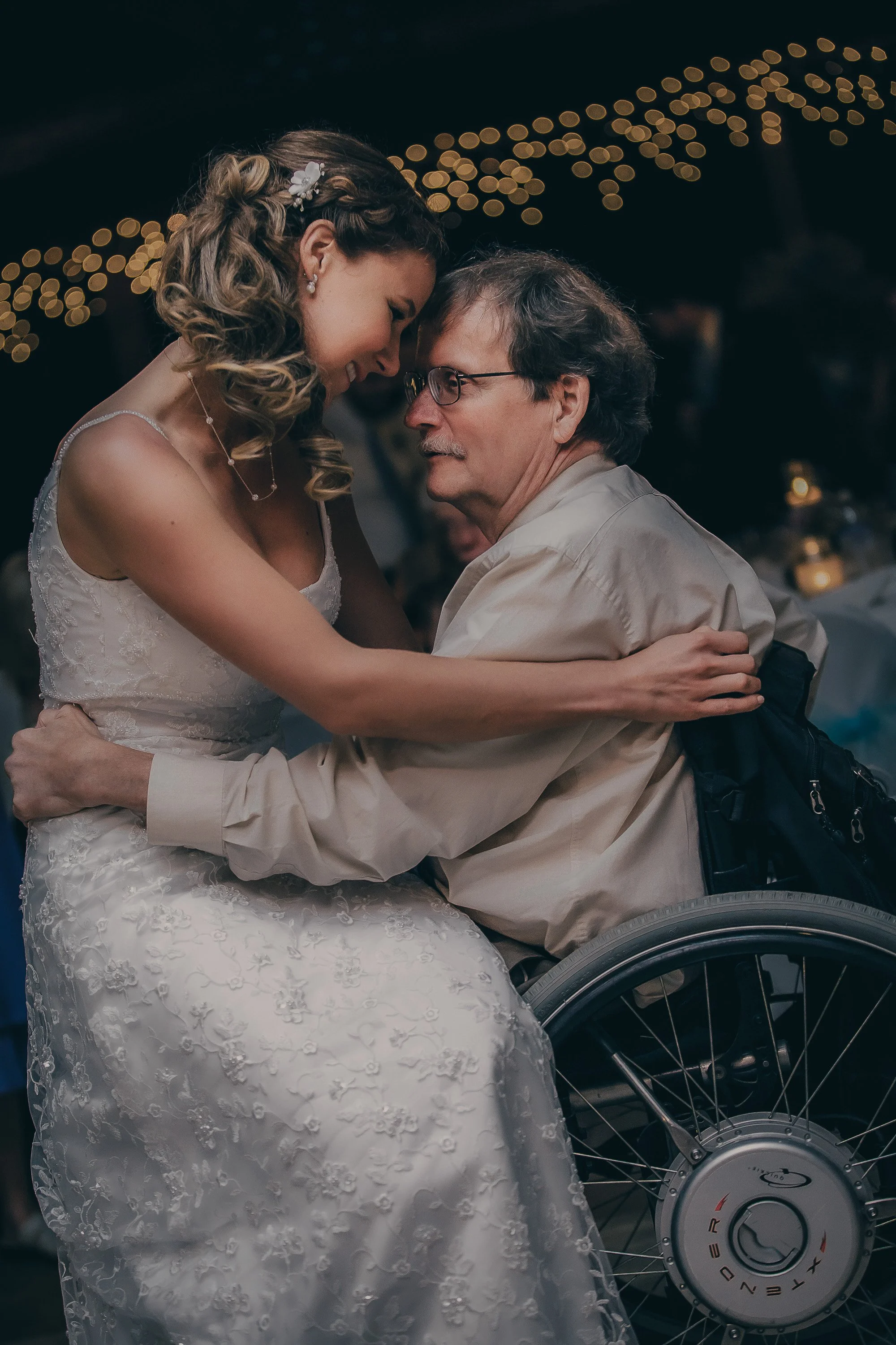 A woman in a wedding dress and a man in a wheelchair sharing a joyful dance at a wedding reception, with string lights in the background.