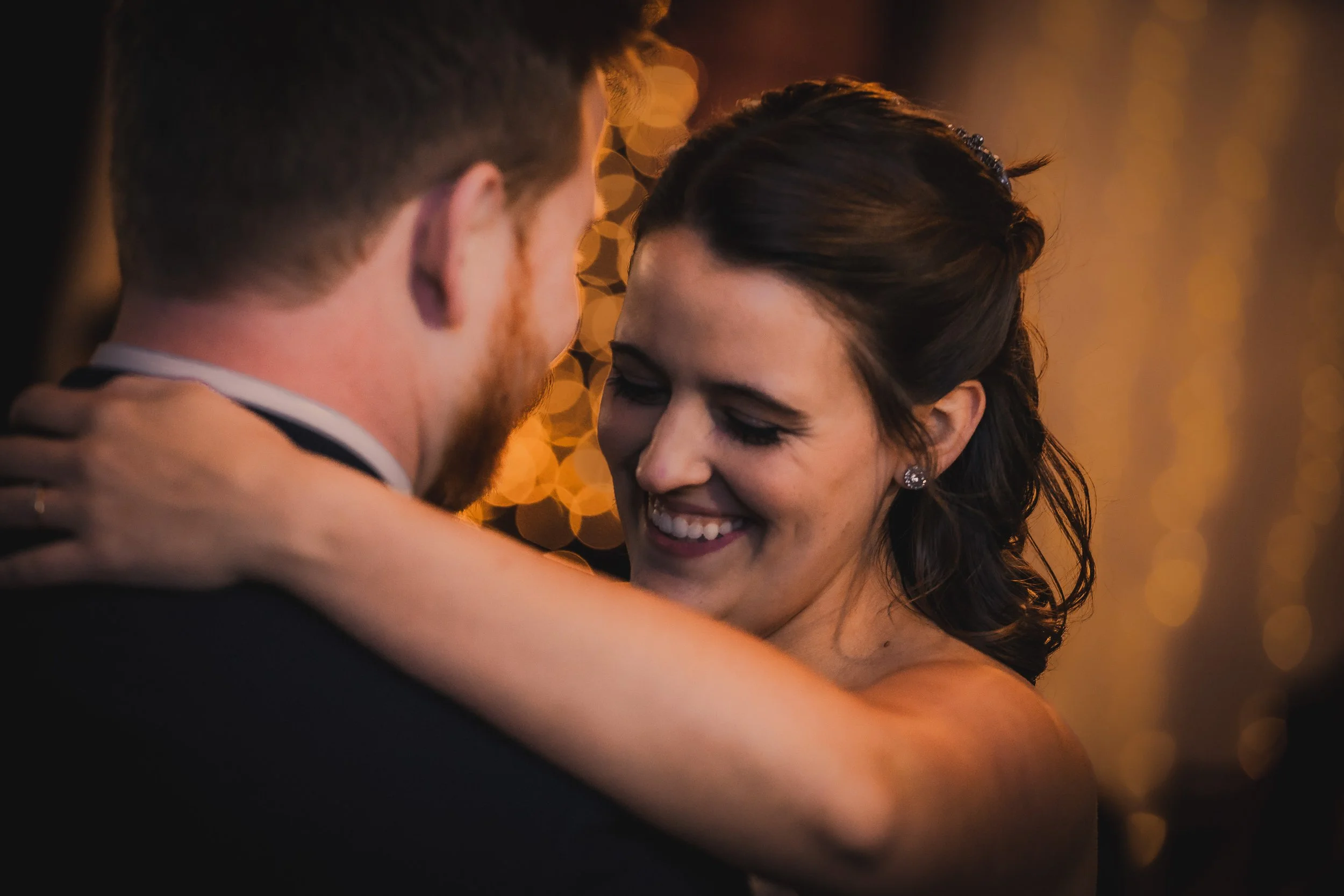 A bride and groom sharing a dance at their wedding, smiling softly at each other with warm, golden lighting in the background.