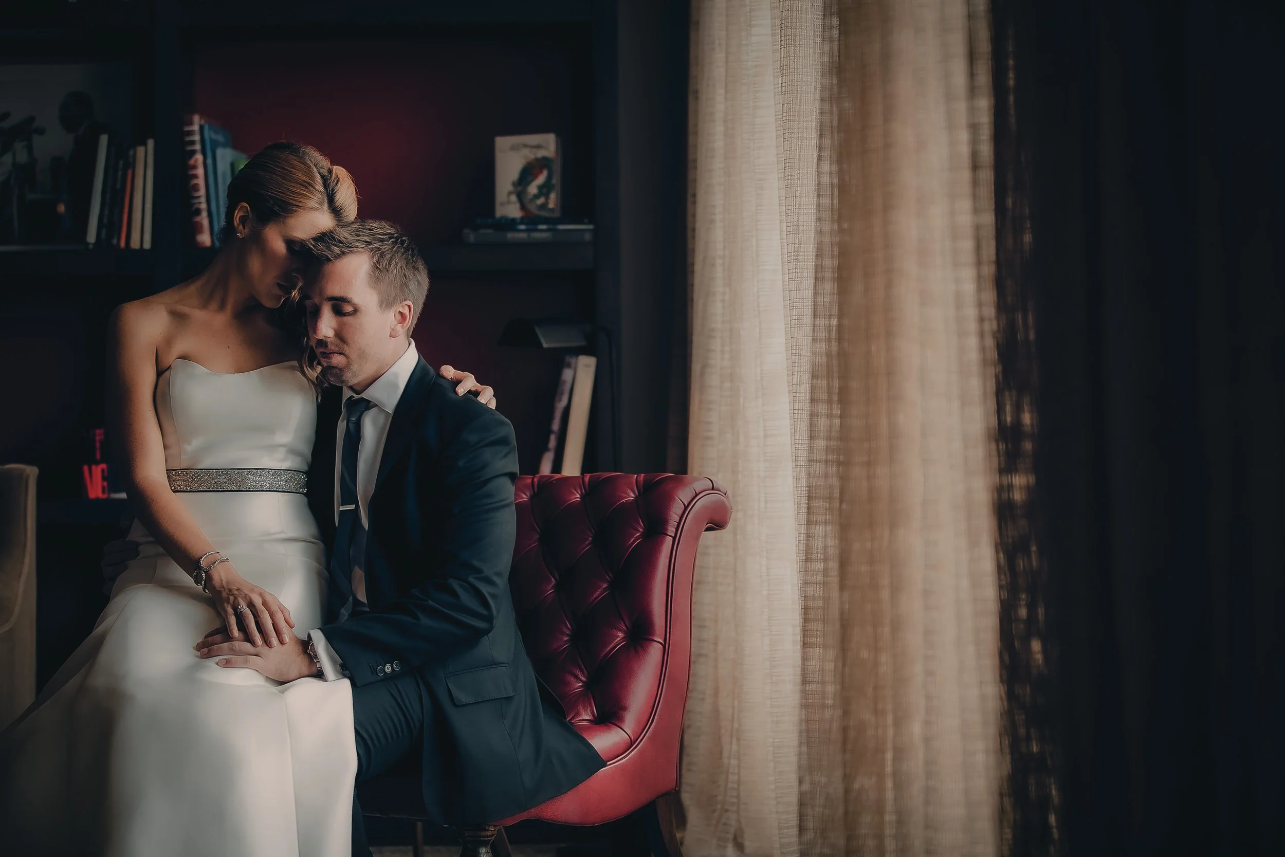 A couple in wedding attire sitting closely on a red armchair, with the woman resting her head on the man's shoulder in a dimly lit room.