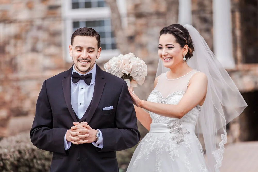 A smiling bride tap's a grooms shoulder from behind during their first look.
