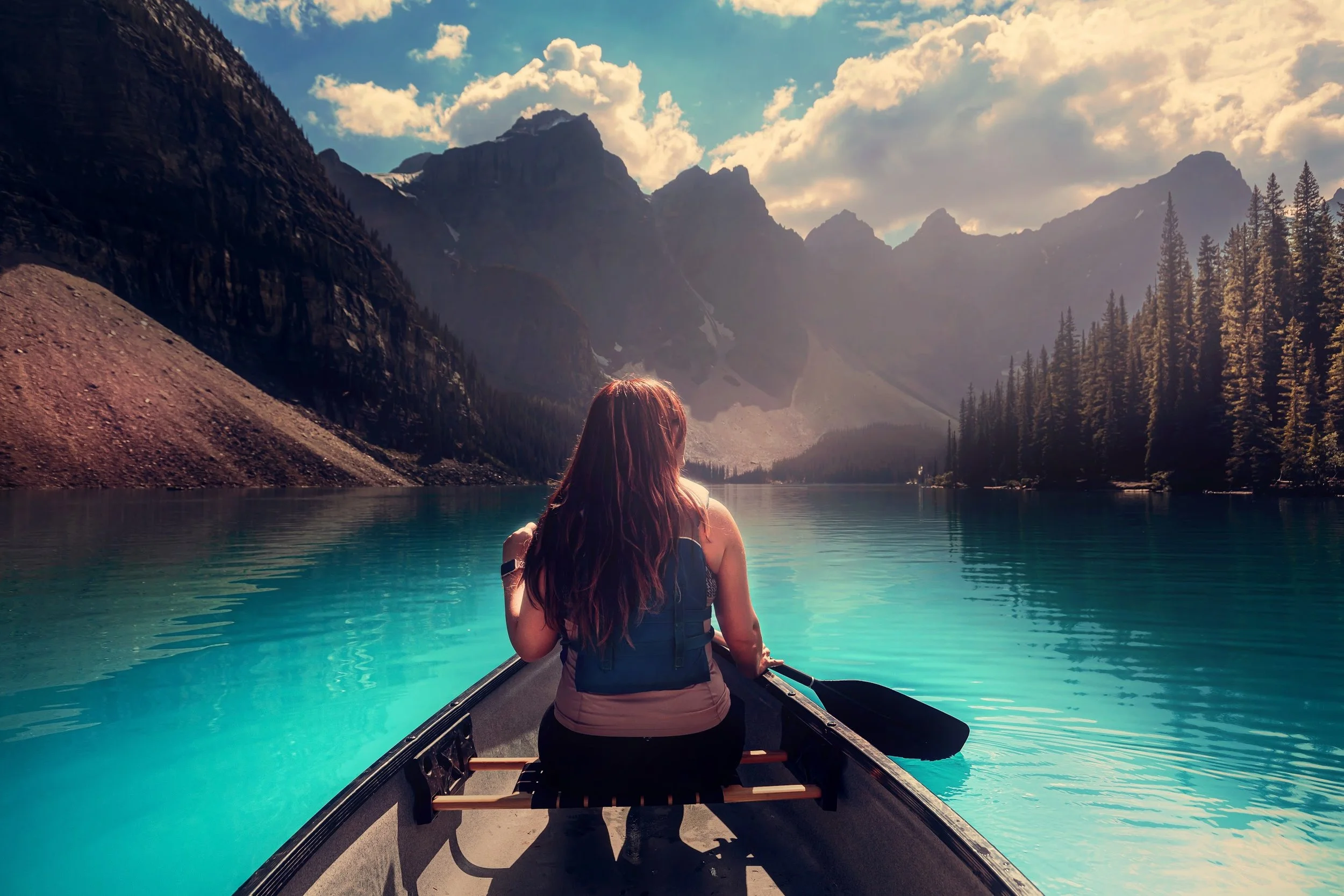 A woman sitting in a kayak on a turquoise lake surrounded by tall mountains and dense evergreen trees, with clouds and blue sky overhead.