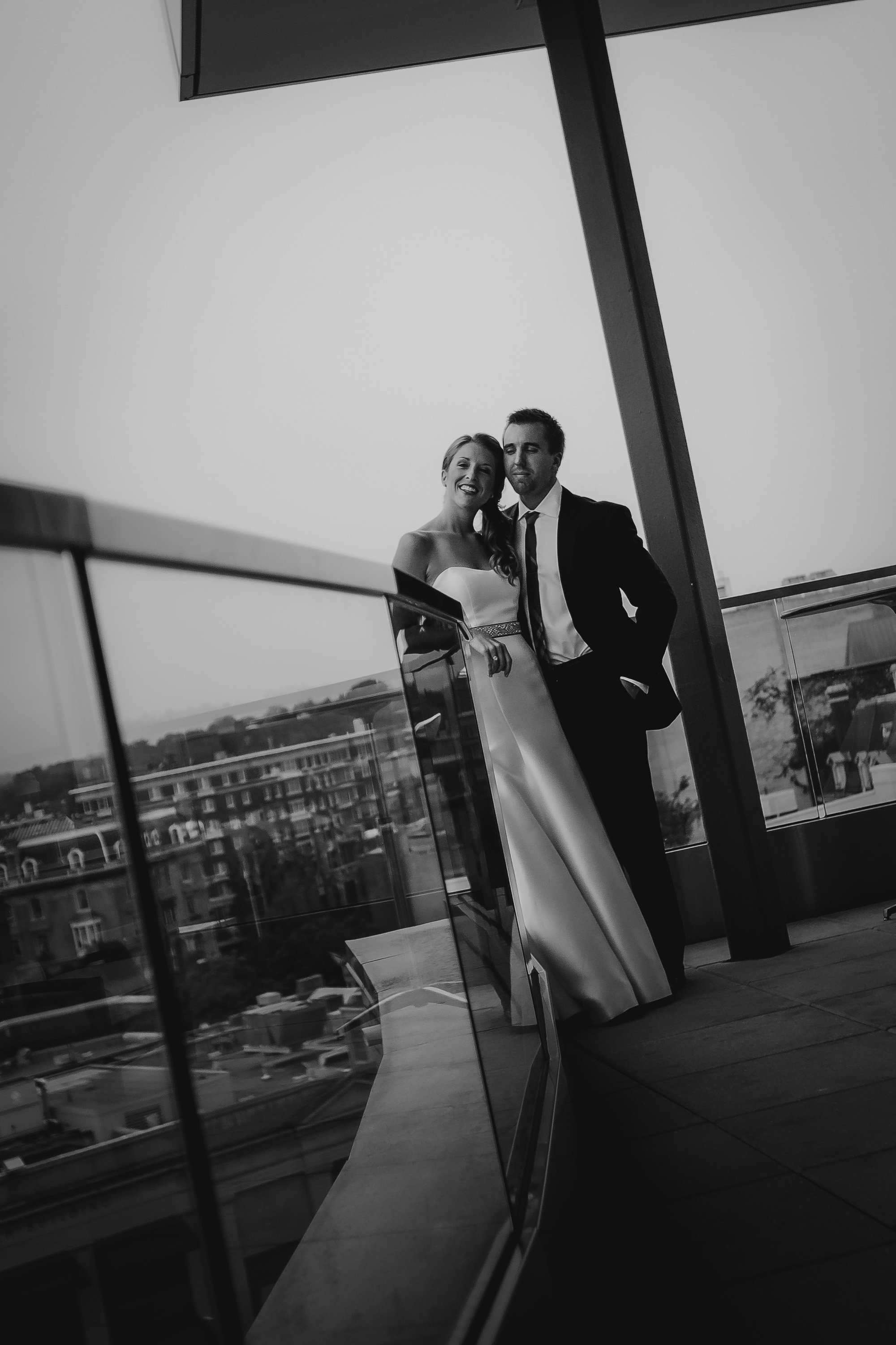Black and white photo of a bride and groom on a balcony, smiling and looking at the camera, with city buildings in the background.