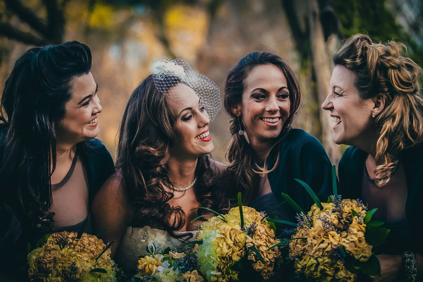 Four women smiling and outdoors, three holding bouquets of yellow flowers, with a woman in wedding attire in the middle wearing a veil, surrounded by trees with fall foliage.