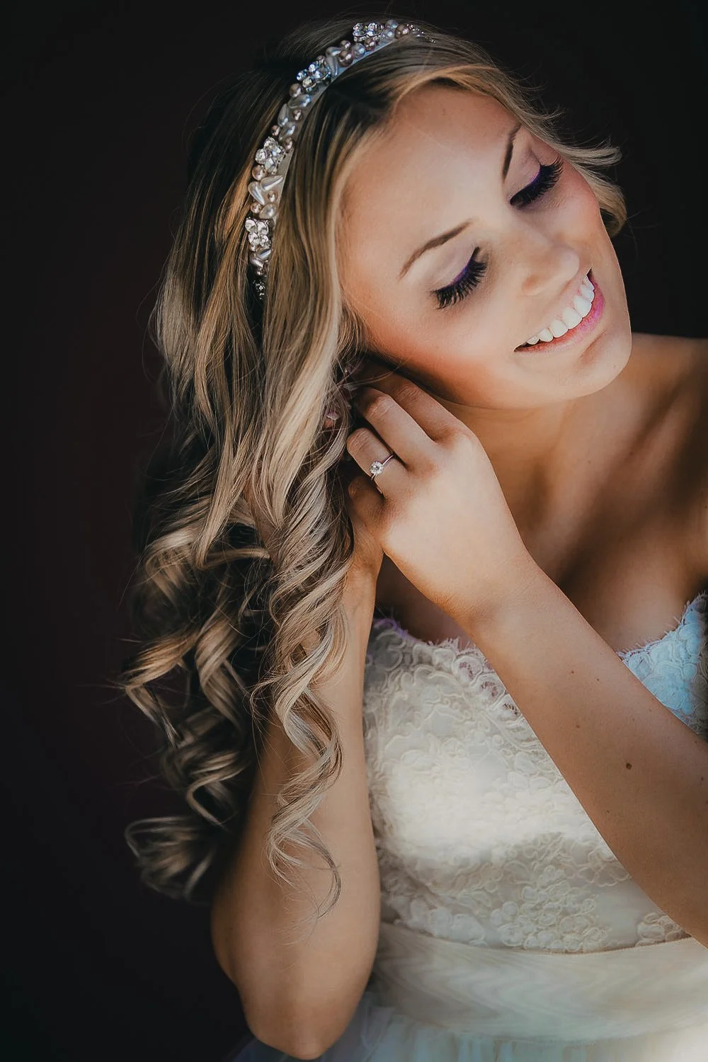 A young woman with blonde wavy hair wearing a pearl and rhinestone headband, earrings, and a white lace dress. She is smiling with closed eyes and touching her ear, showing a ring on her finger.