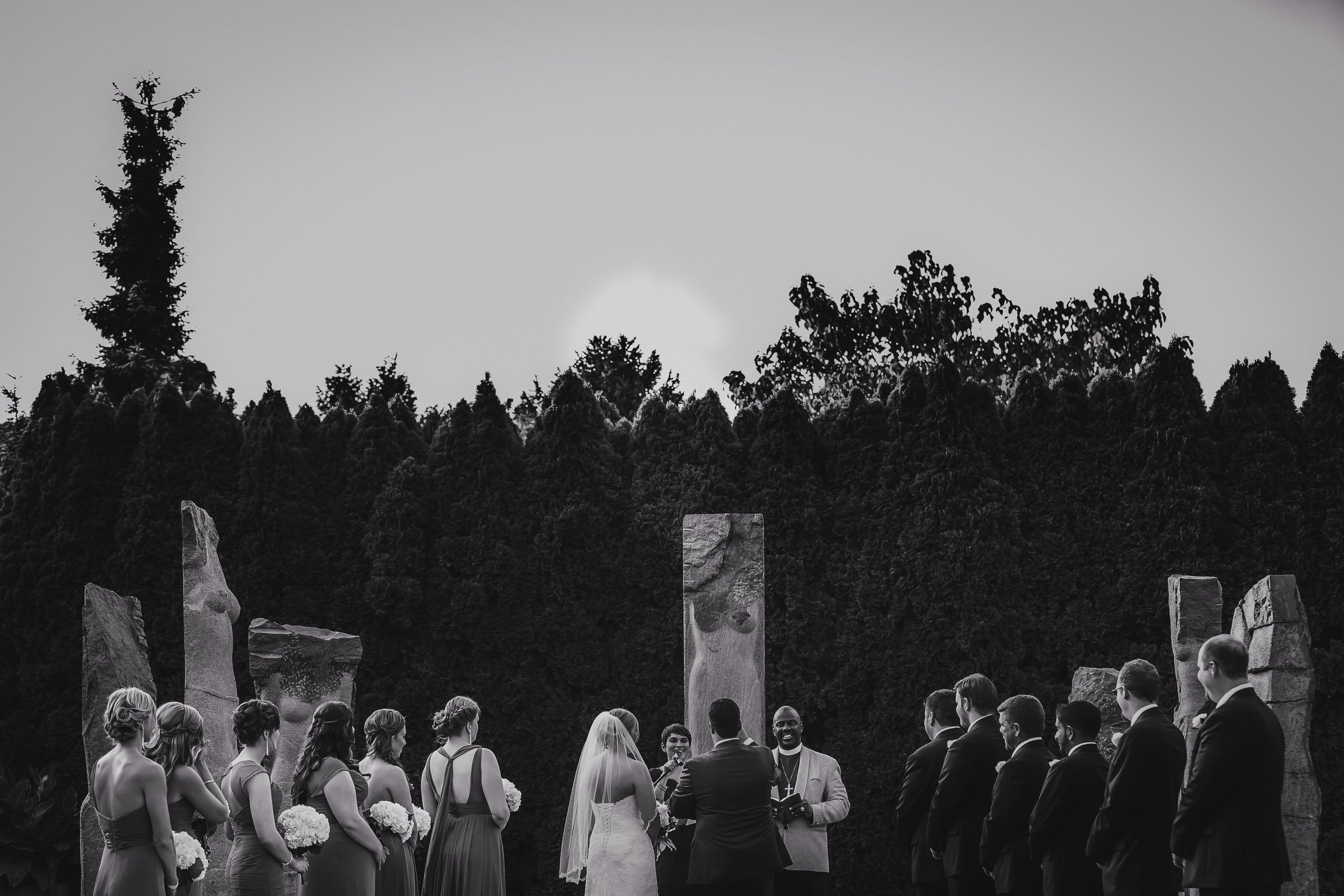 A black and white outdoor wedding ceremony with the bride and groom exchanging vows, surrounded by bridesmaids and groomsmen, amid rock formations and trees.