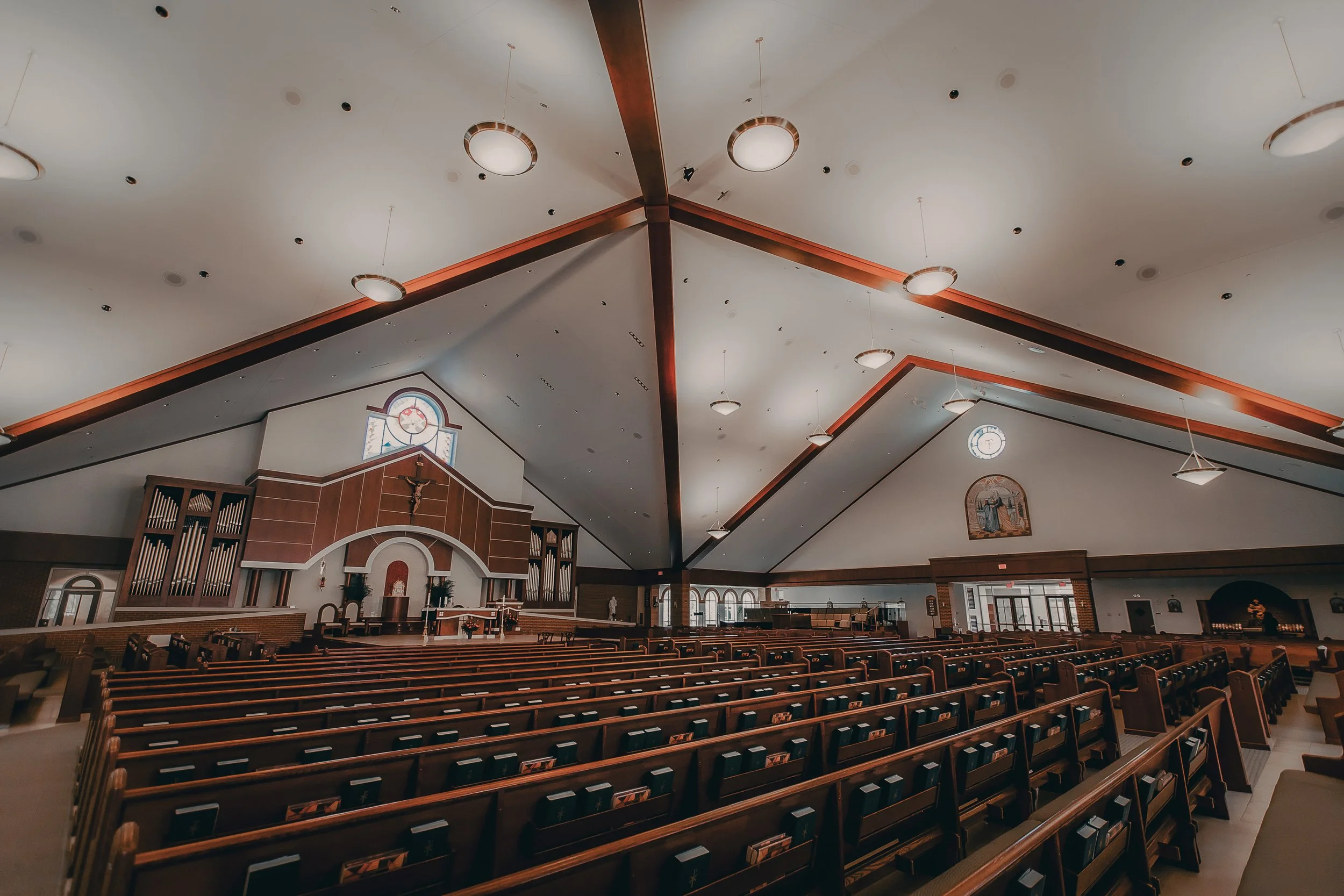 Interior of a church with rows of wooden pews, a raised choir loft, and religious artwork, with high vaulted ceilings and hanging circular light fixtures.