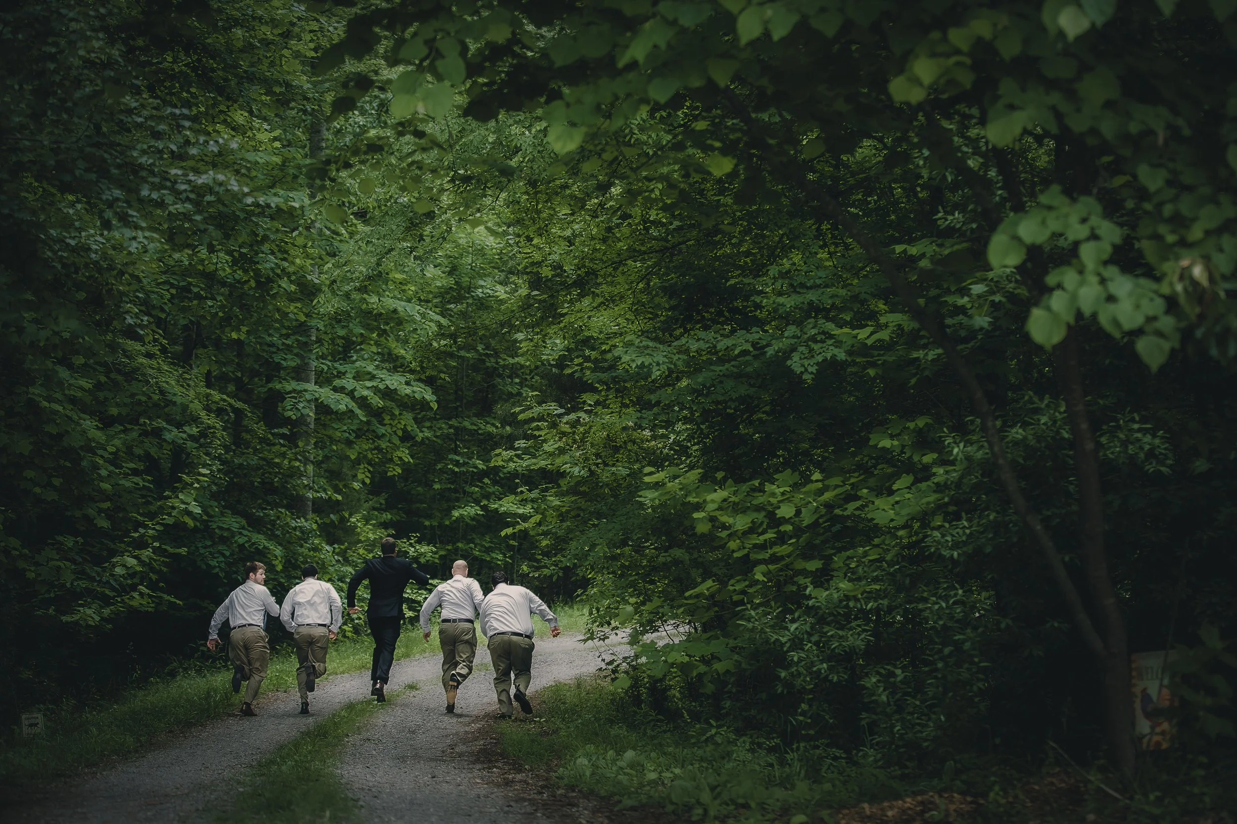 A group of five men running on a forest trail surrounded by green trees and foliage.