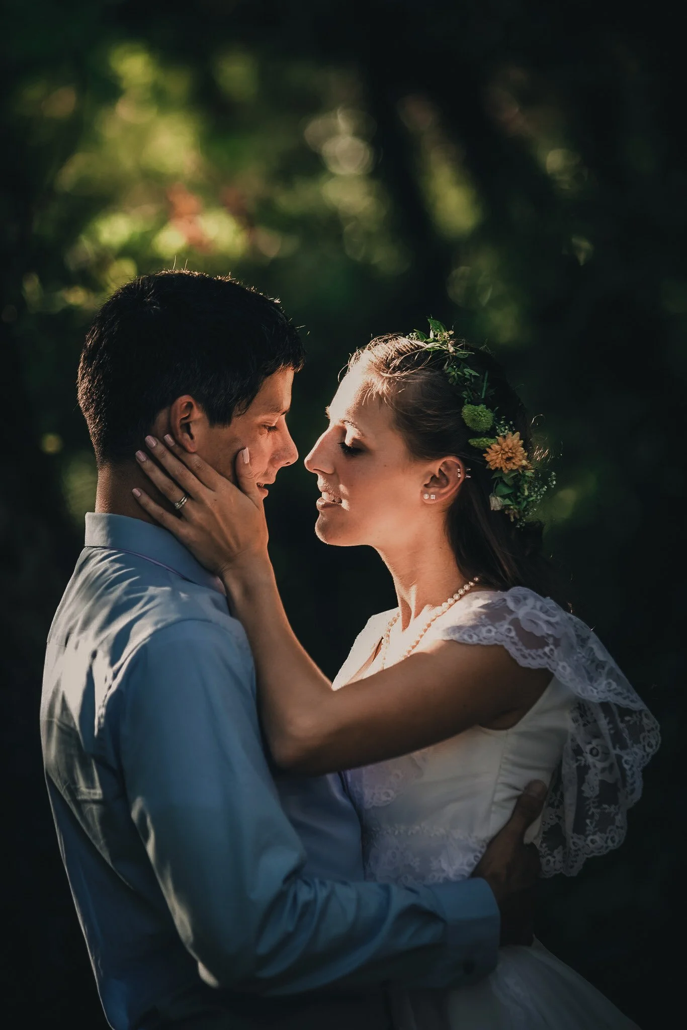 A couple in wedding attire embracing outdoors with sunlight filtering through trees in the background.