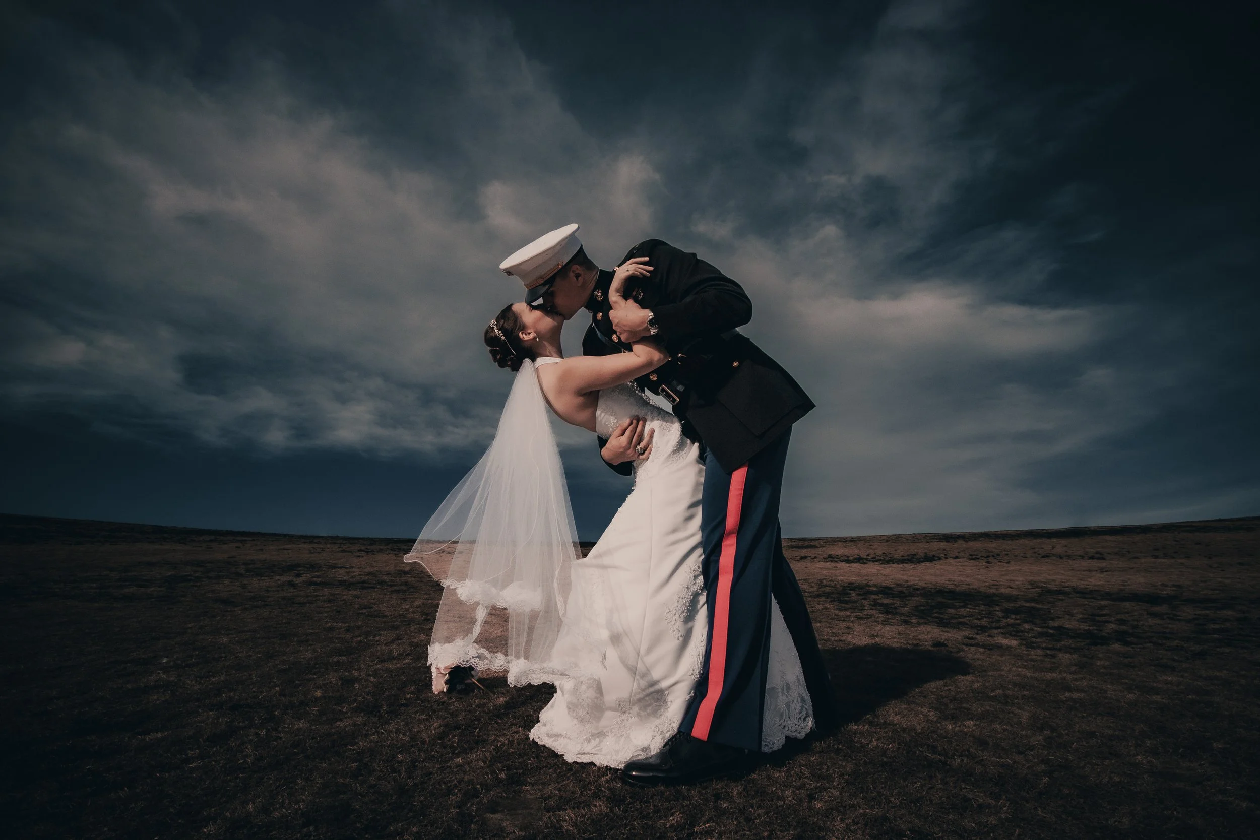 A couple in wedding attire sharing a kiss outdoors under a cloudy sky. The woman is wearing a white wedding dress with a veil, and the man is in a military uniform.