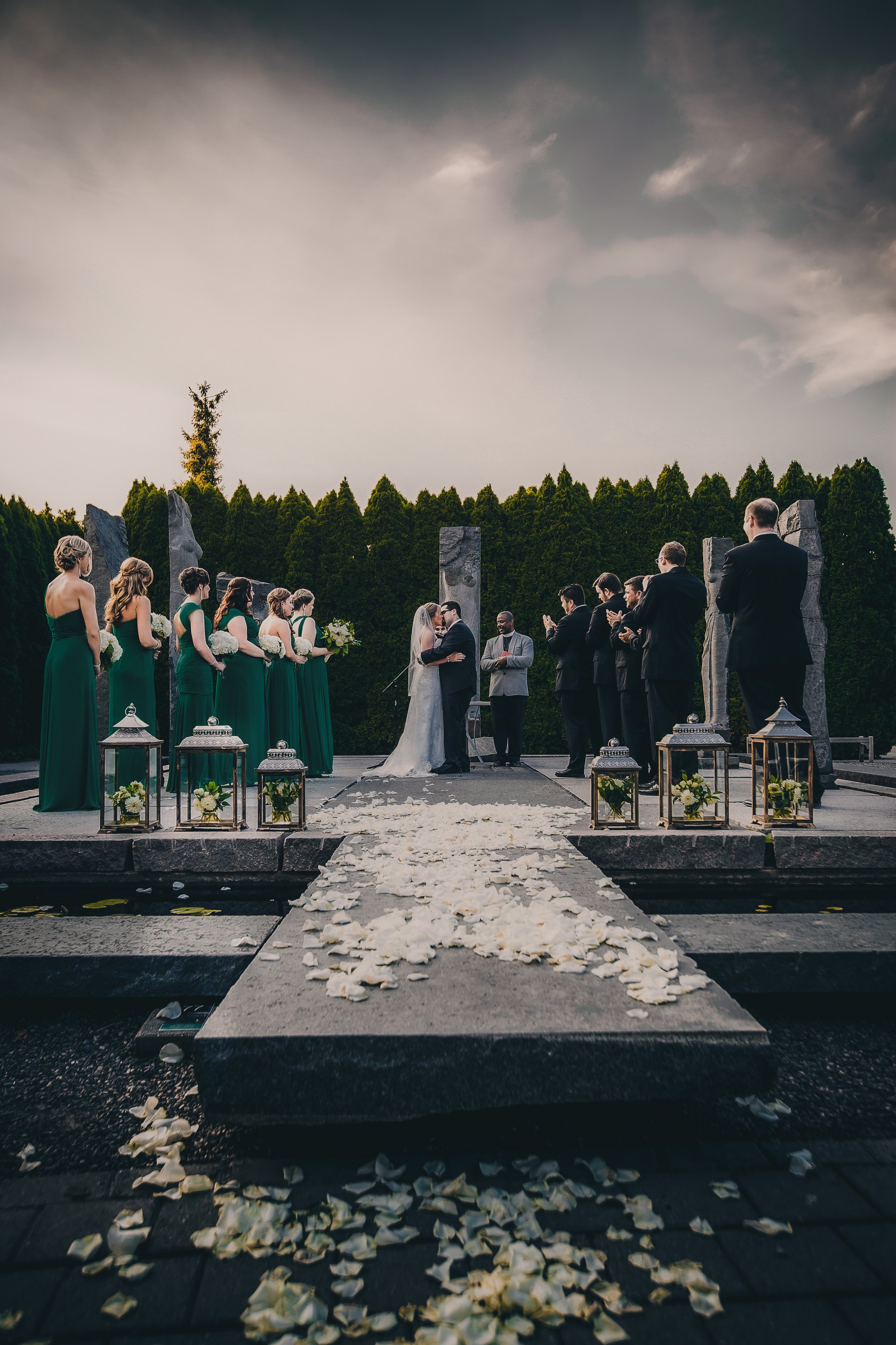 A wedding ceremony taking place outdoors with a bride and groom kissing at the altar, surrounded by bridesmaids in green dresses holding bouquets, groomsmen in suits, and officiants. Petals are scattered along the aisle with lantern decorations on ei