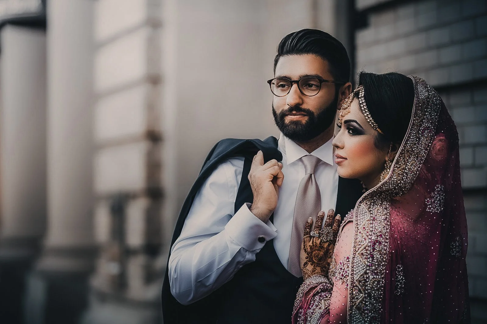 A man and woman standing close together, the man in a suit and tie with glasses carrying a backpack over his shoulder, and the woman dressed in traditional South Asian bridal attire with ornate jewelry and henna on her hands, standing outdoors agains