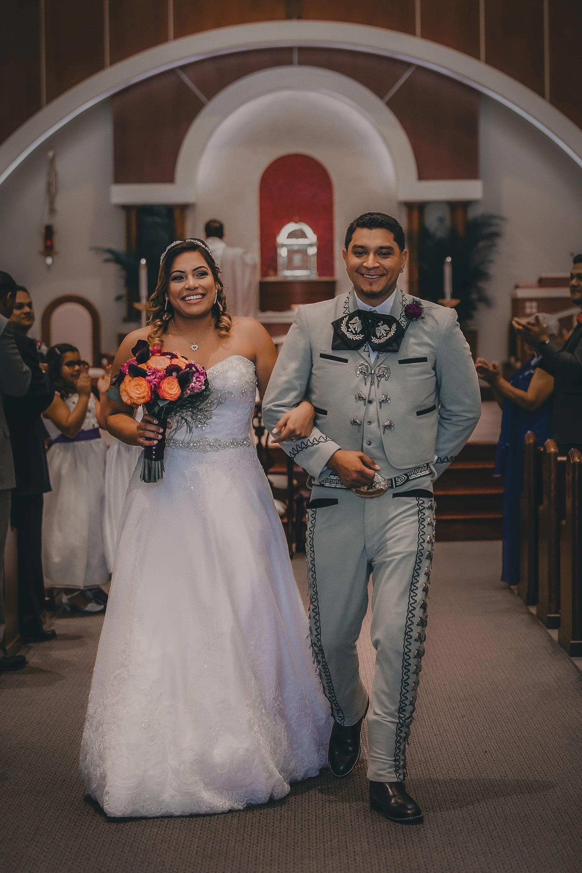A bride in a white wedding dress holding a bouquet and a groom in traditional Mexican mariachi attire walking down the aisle in a church, surrounded by guests.