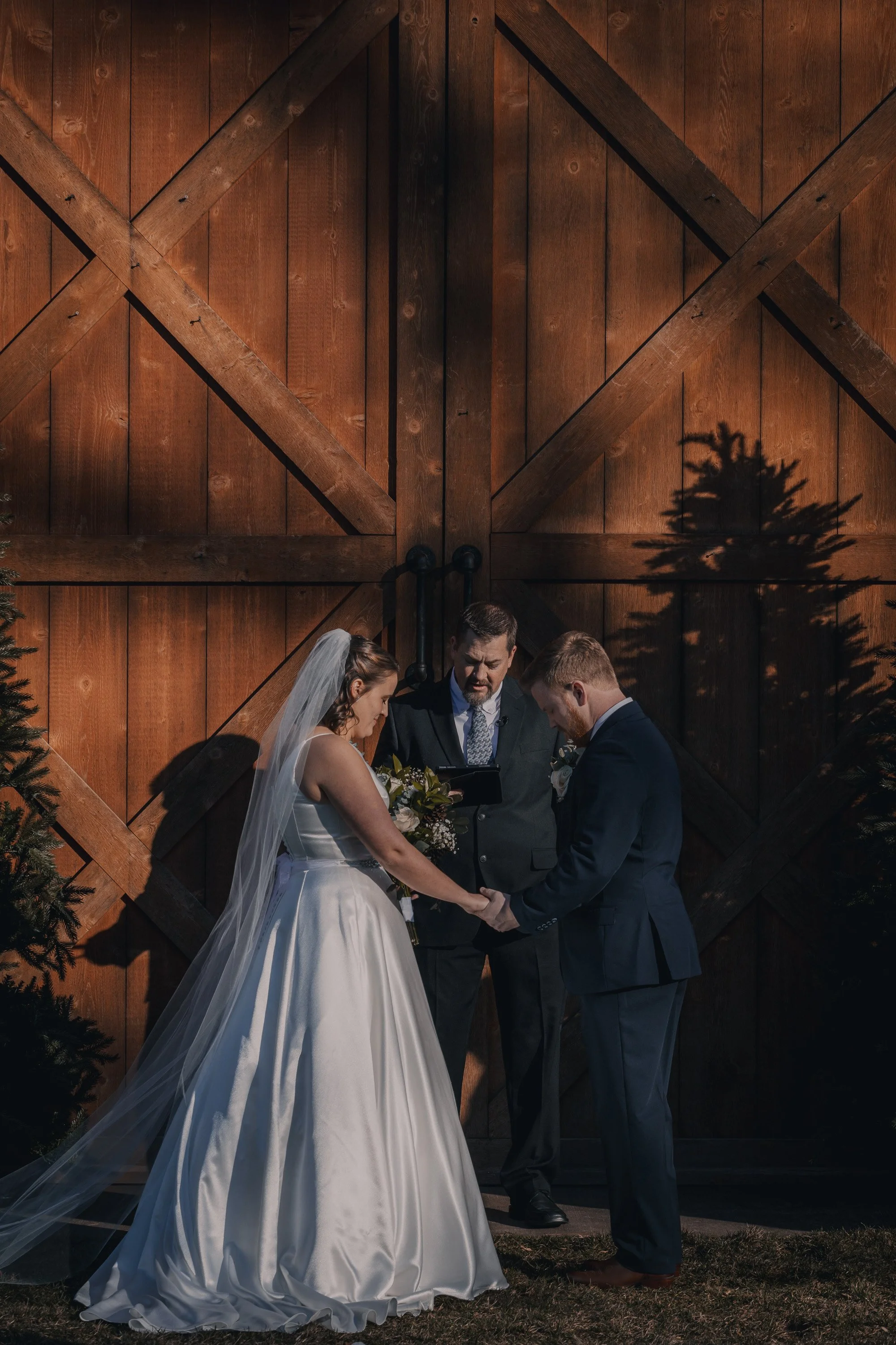 A bride and groom holding hands during their wedding ceremony outdoors, with an officiant reading vows, against a large wooden barn door with shadows of trees.