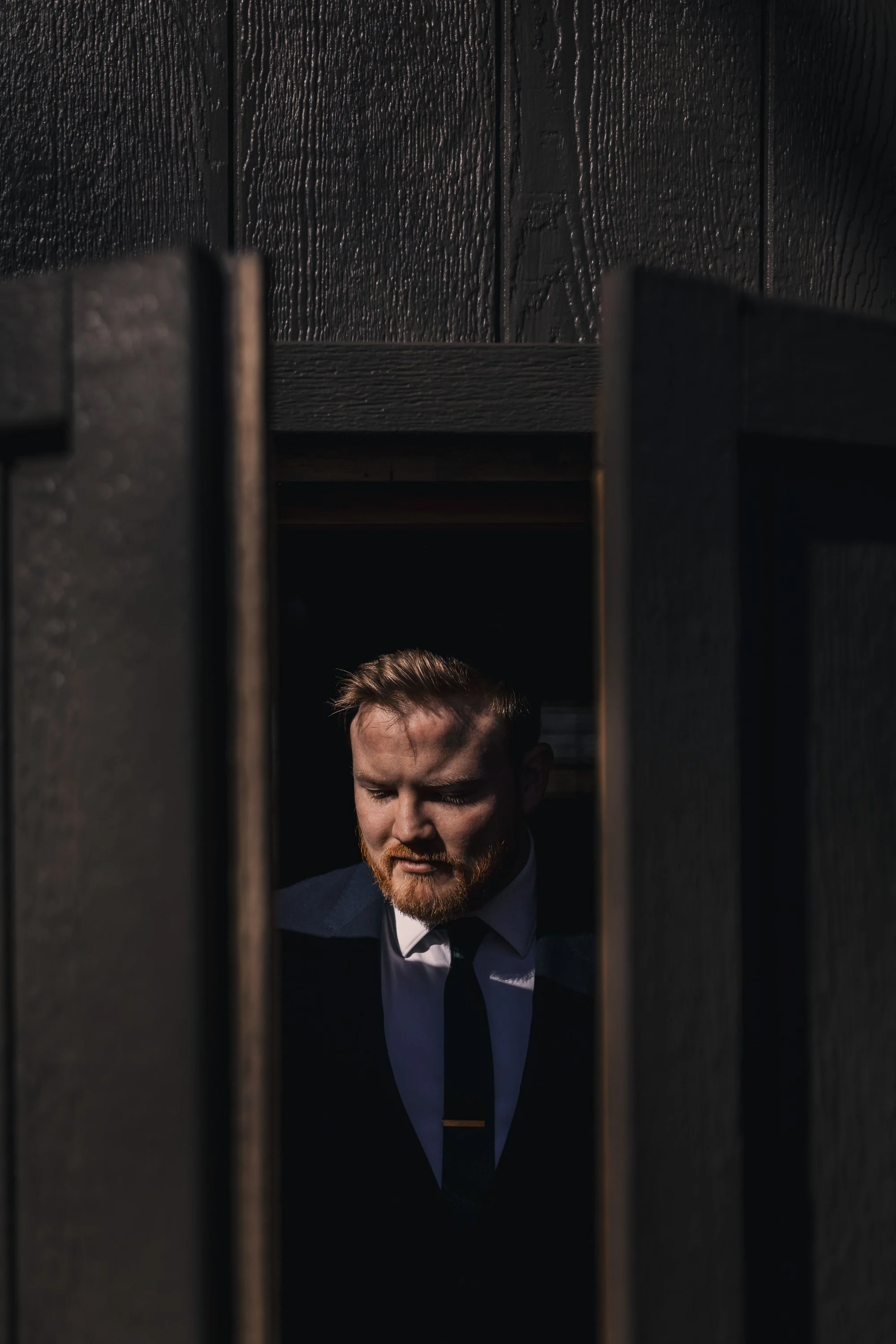 A man in a dark suit and tie looking downward, seen through a narrow gap between dark wooden doors in a dimly lit setting.