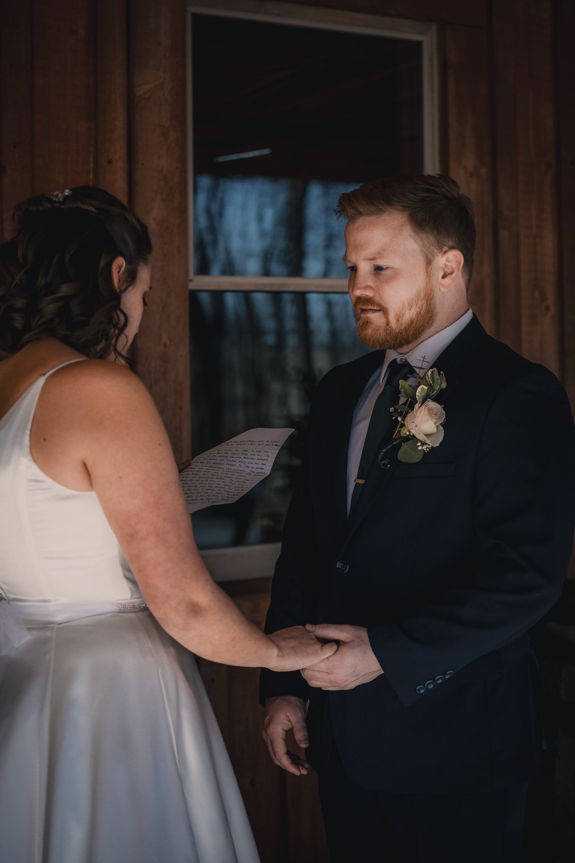 A bride and groom exchange vows indoors, holding hands, with the bride reading from a paper, against a wooden wall background.