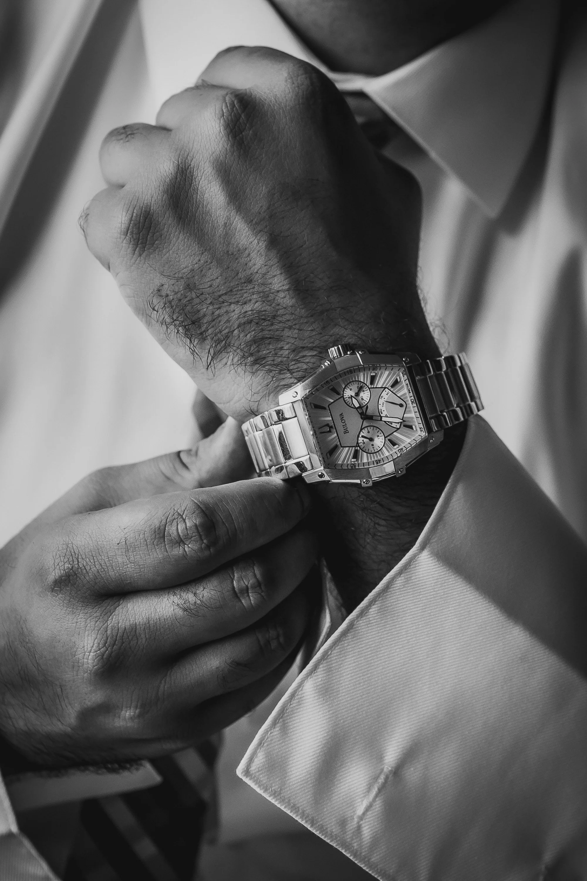 A man adjusting his wristwatch with a metal band while wearing a light-colored suit and tie.