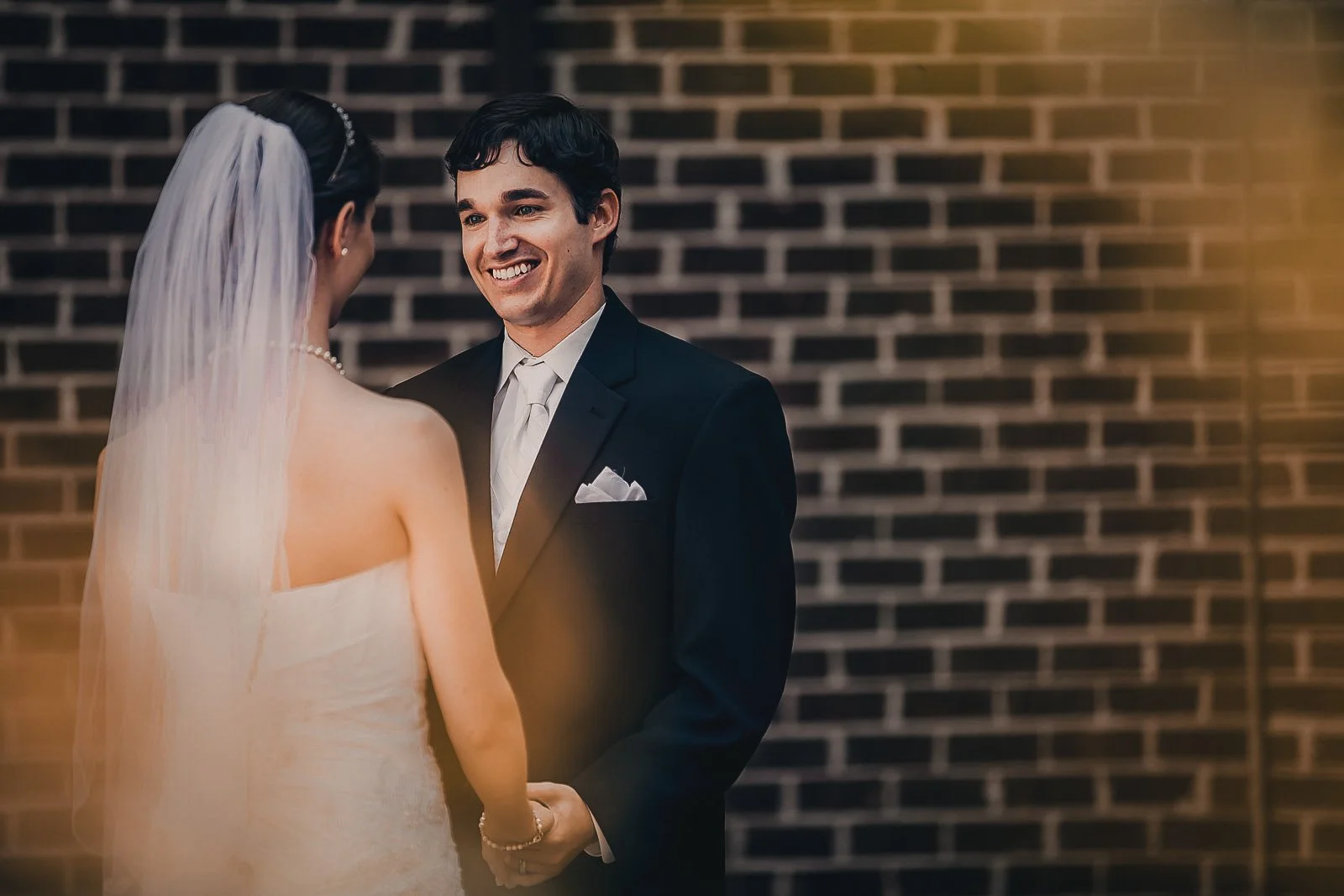 A bride and groom holding hands, facing each other during their wedding ceremony, with a brick wall in the background.