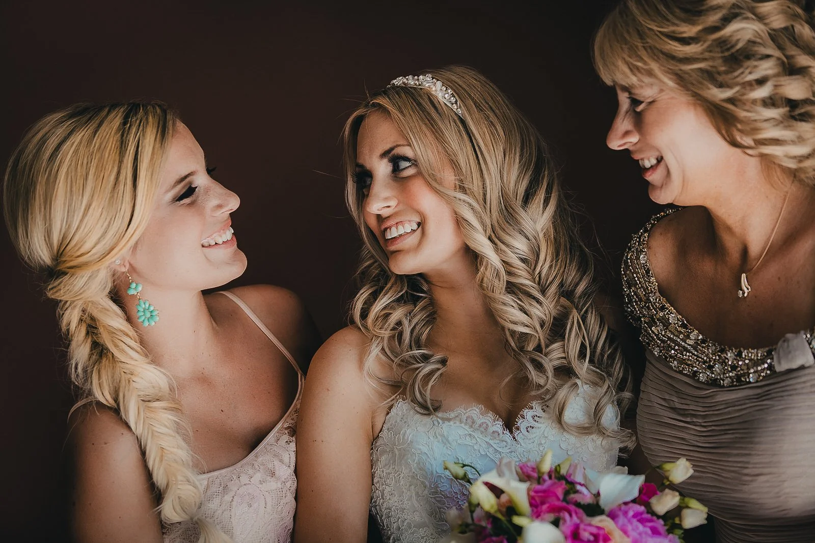 A bride with wavy blonde hair holding a bouquet, smiling and looking at two women, one with long blonde hair and the other with curly blonde hair, dressed in elegant outfits, smiling.