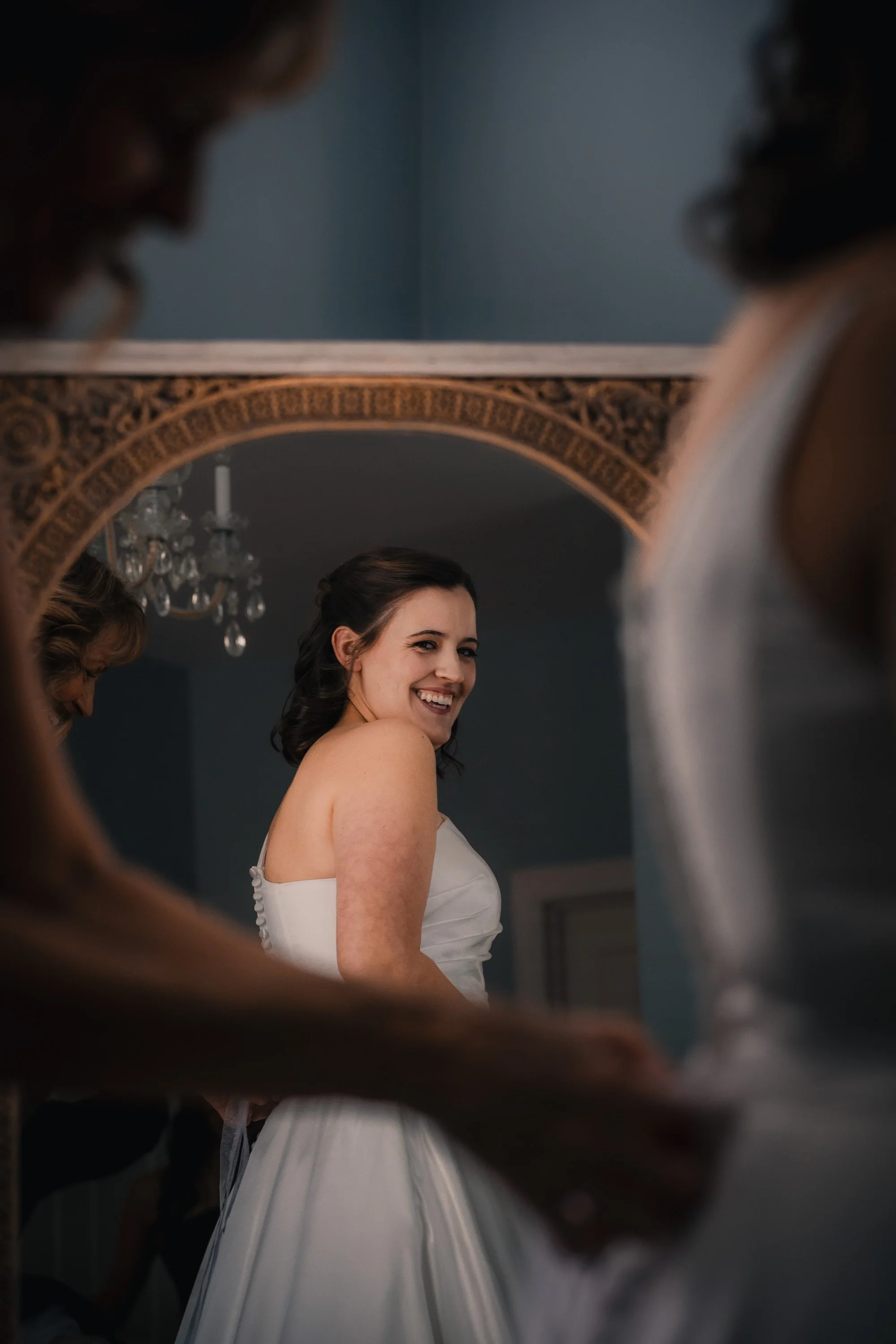 A bride smiling in front of a mirror, wearing a strapless white wedding dress, with a chandelier visible in the background.