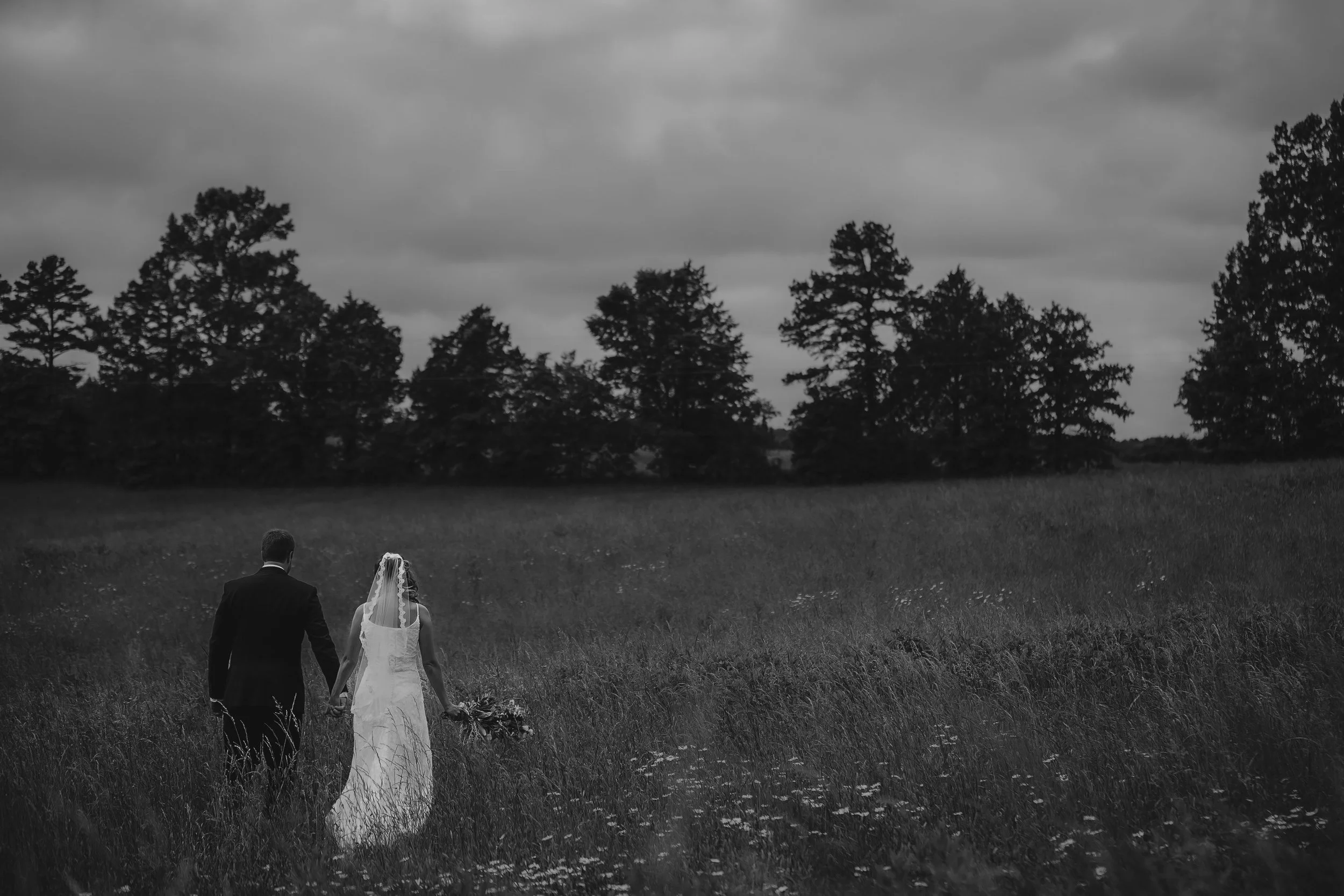 A bride and groom walking hand in hand through a grassy field with trees in the background on a cloudy day.