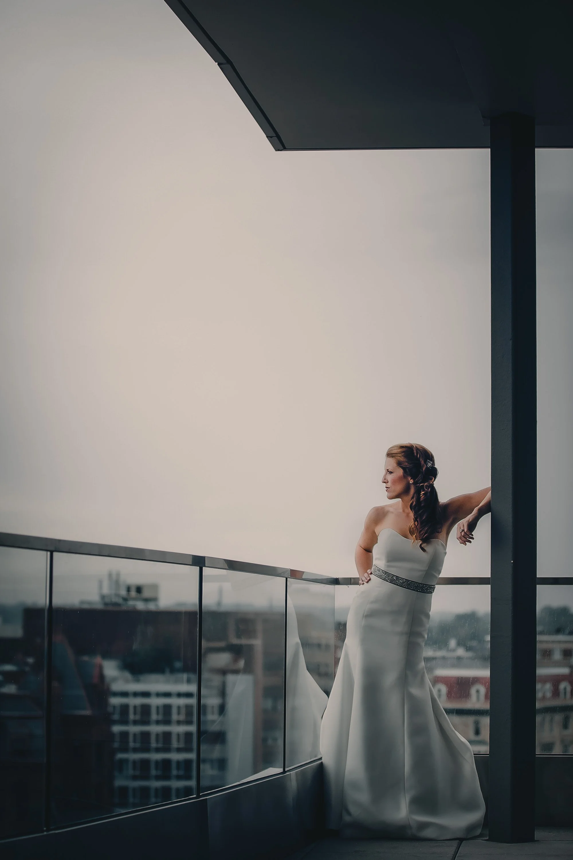 A woman in a strapless white gown with a bejeweled belt, leaning on a balcony railing on a rooftop, gazing into the distance with city buildings in the background.