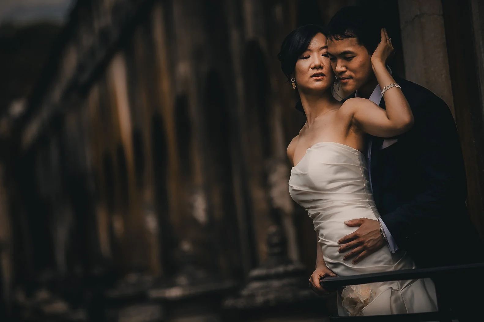 A couple in formal attire close together, the woman in a strapless white dress and the man in a black suit, standing against a dark background.