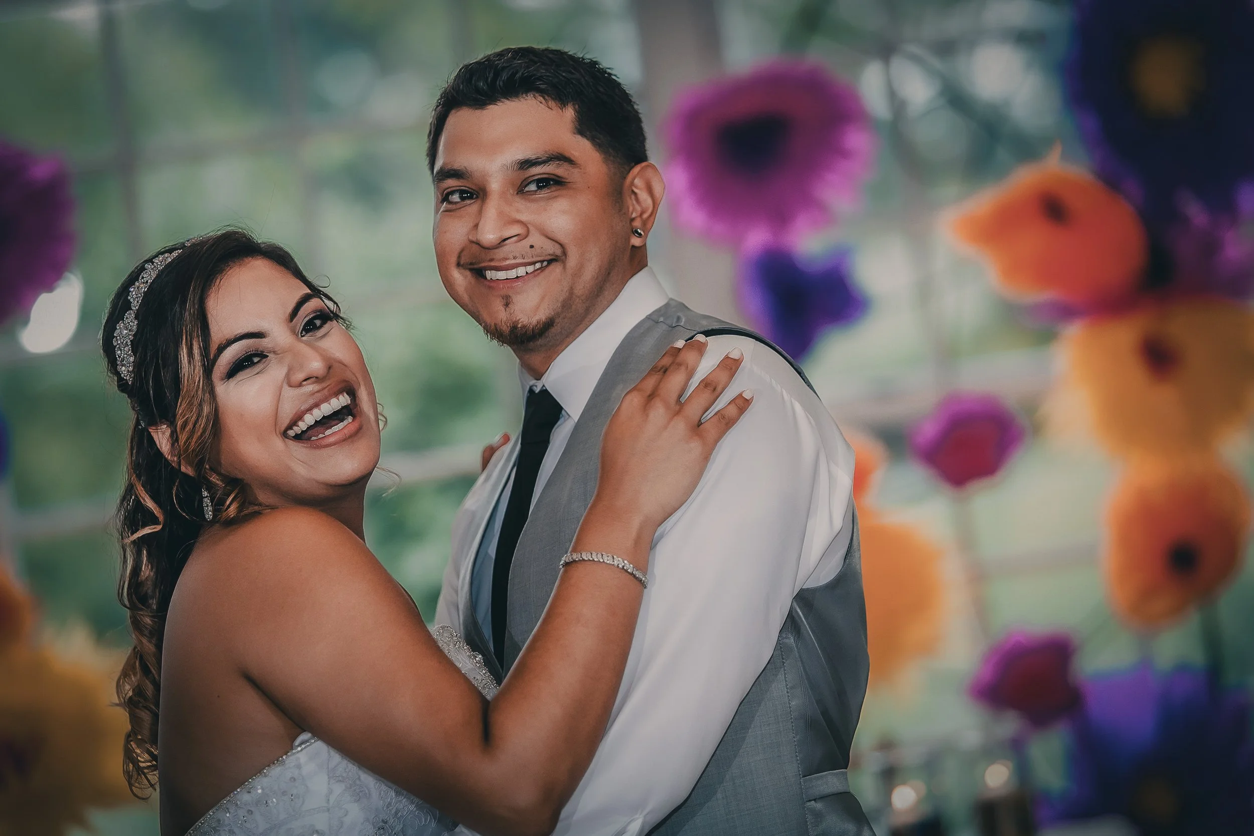 A happy bride and groom dancing at their wedding reception, smiling and embracing, with colorful paper flowers in the background.