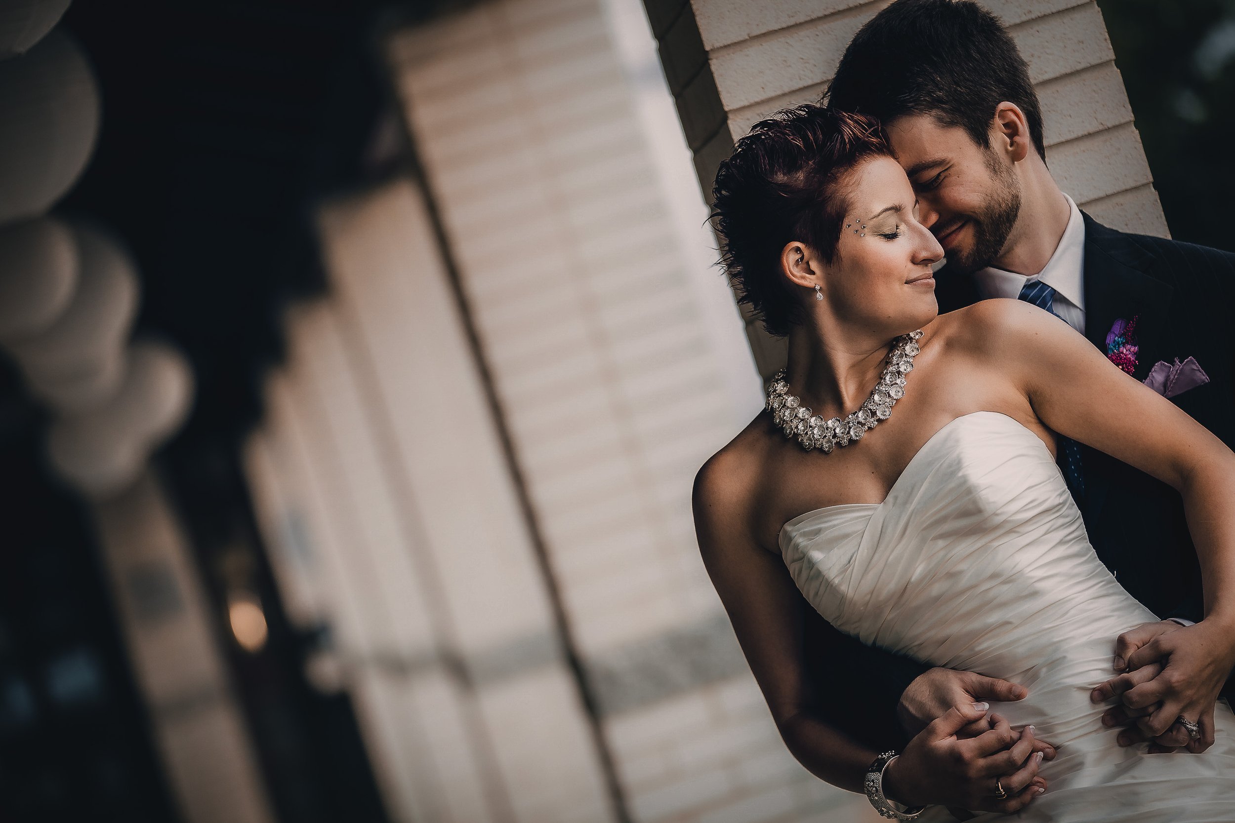 A bride and groom leaning close together, smiling with eyes closed, posing indoors near a brick wall and window.