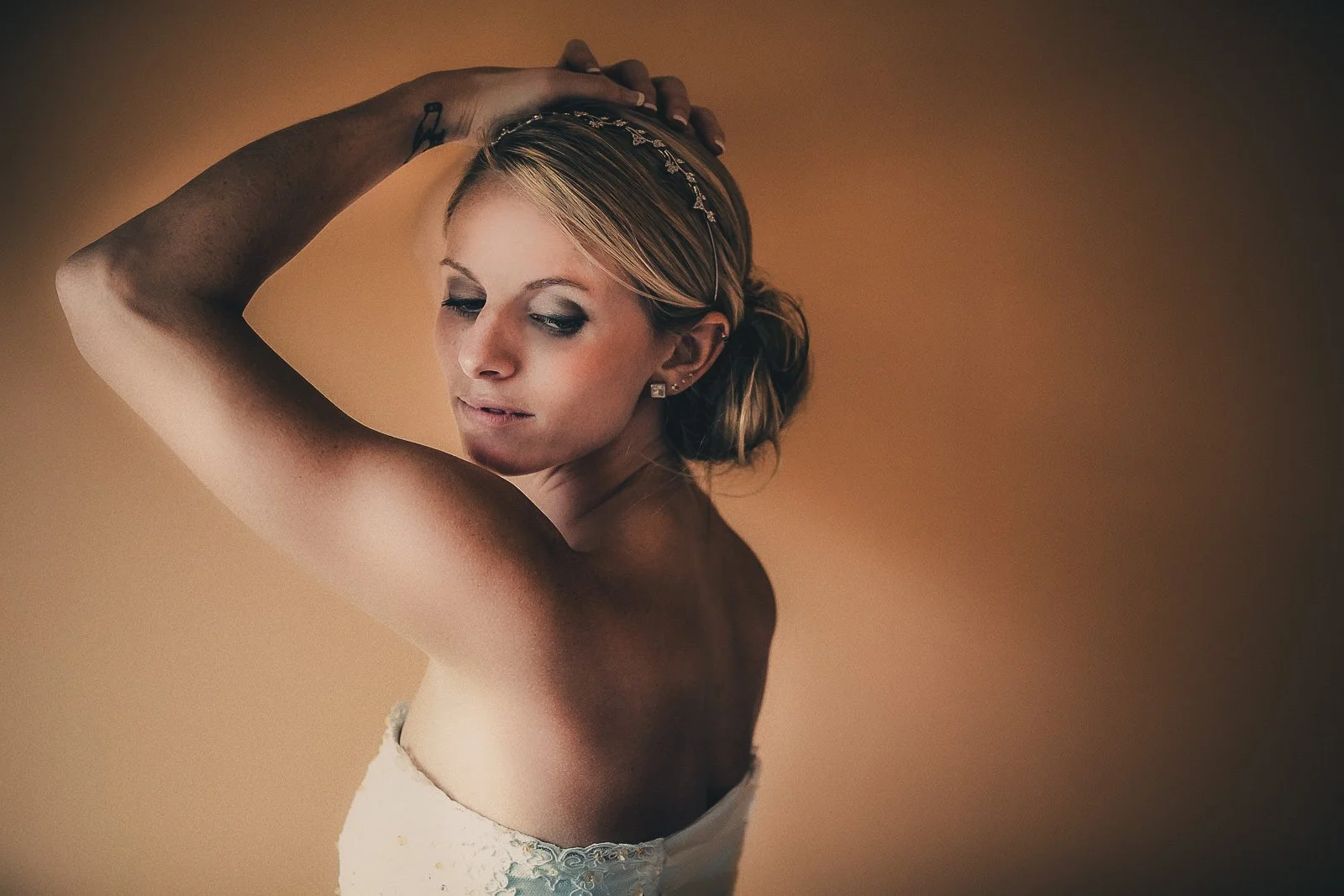 A young woman with blonde hair styled in an updo, wearing earrings and a white strapless dress, posed against a plain brown background, with one arm raised over her head.