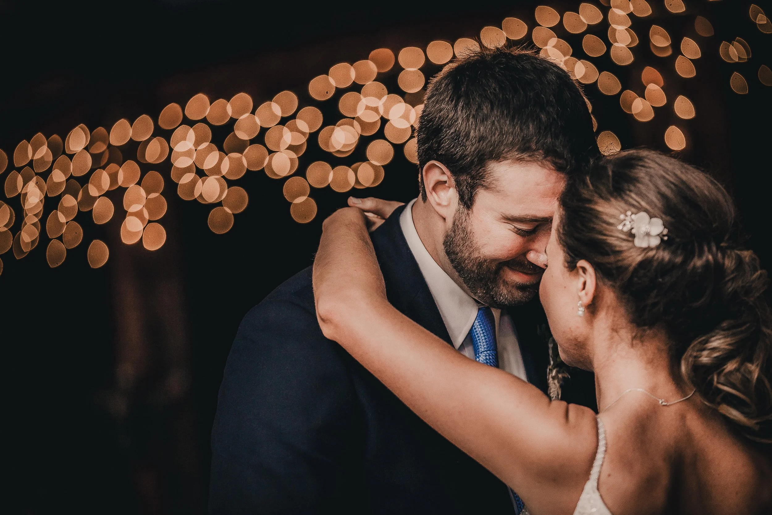A couple in wedding attire sharing a tender moment, with their foreheads touching and eyes closed, against a background of bokeh lights.