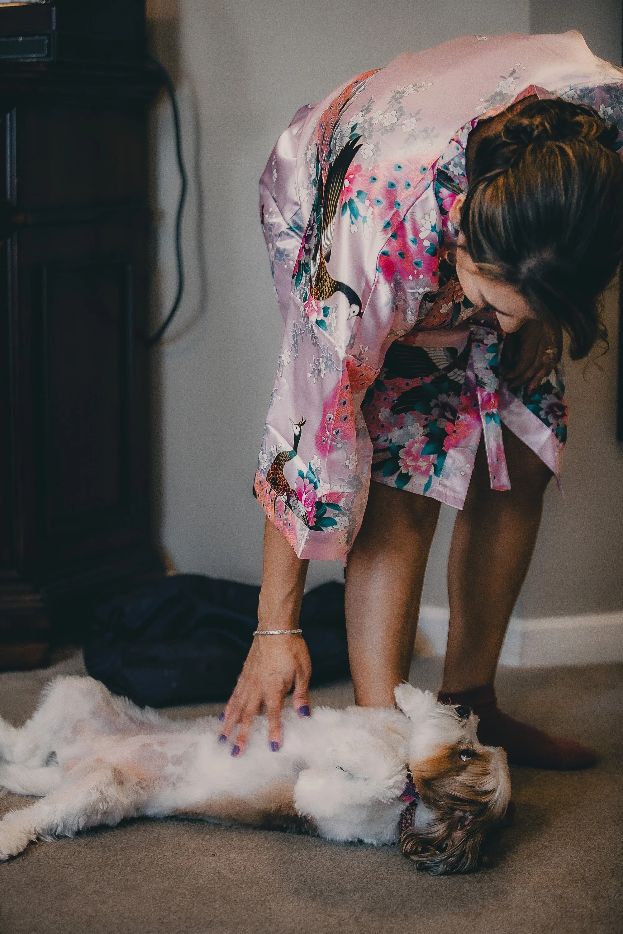 A woman in a pink kimono with a floral and bird pattern is petting a small, white and brown dog lying on its back on the carpet.