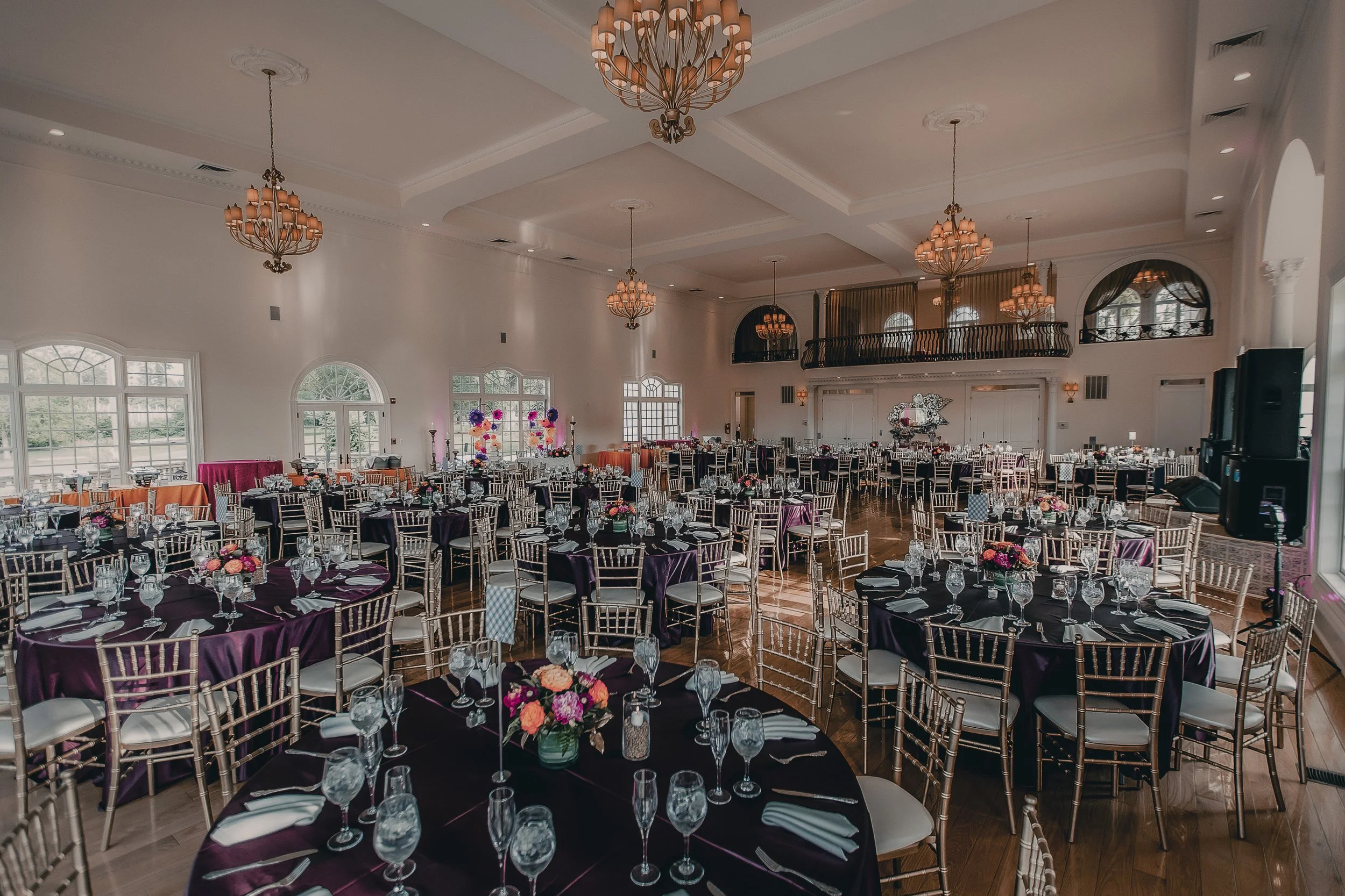 A spacious banquet hall decorated for an event with round tables covered in purple tablecloths, gold chairs, and floral centerpieces. The hall features high ceilings with elegant chandeliers, large windows, and a balcony area at the back.
