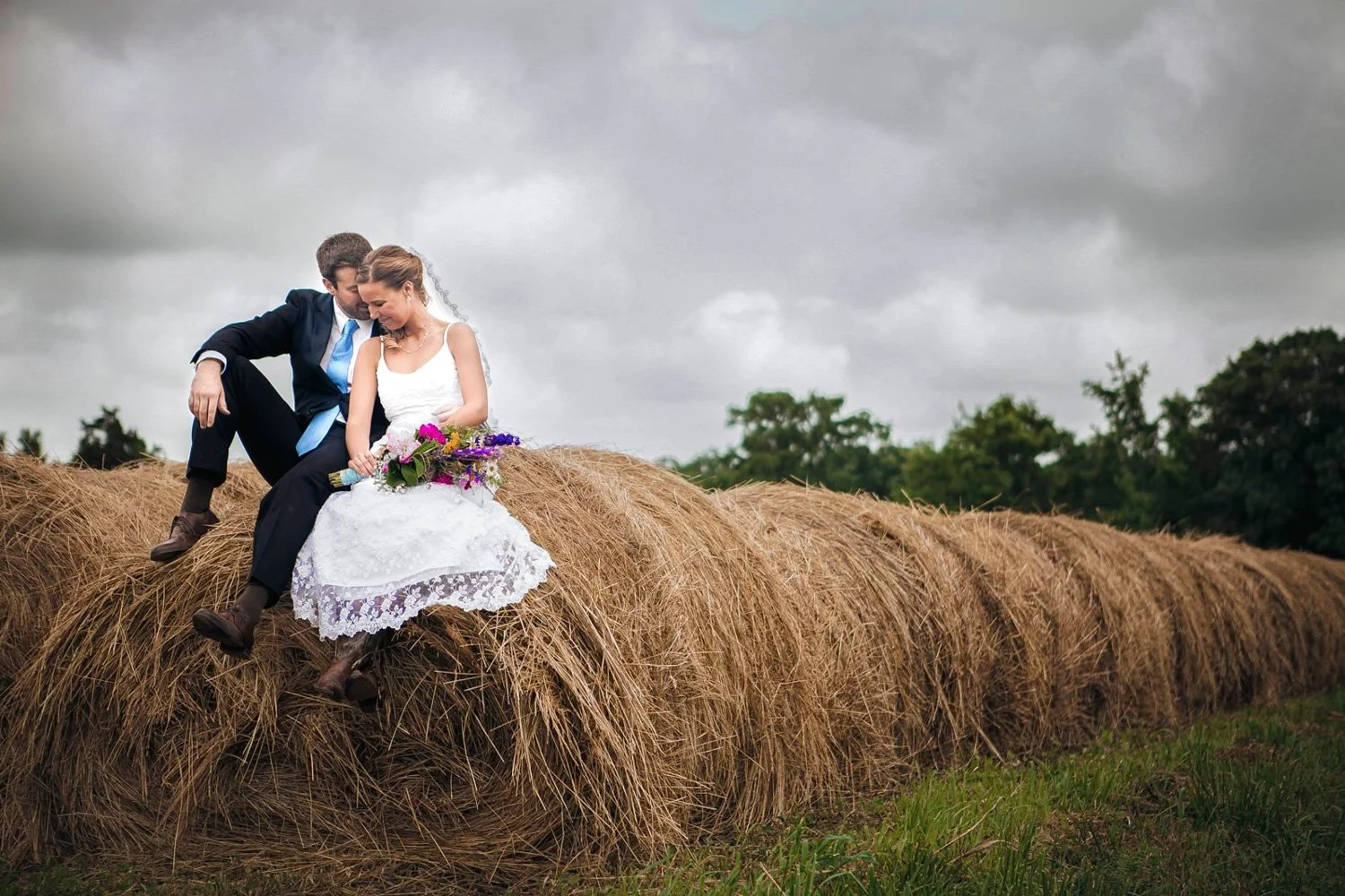 A bride and groom sitting together on top of the end of a row of hay bales.