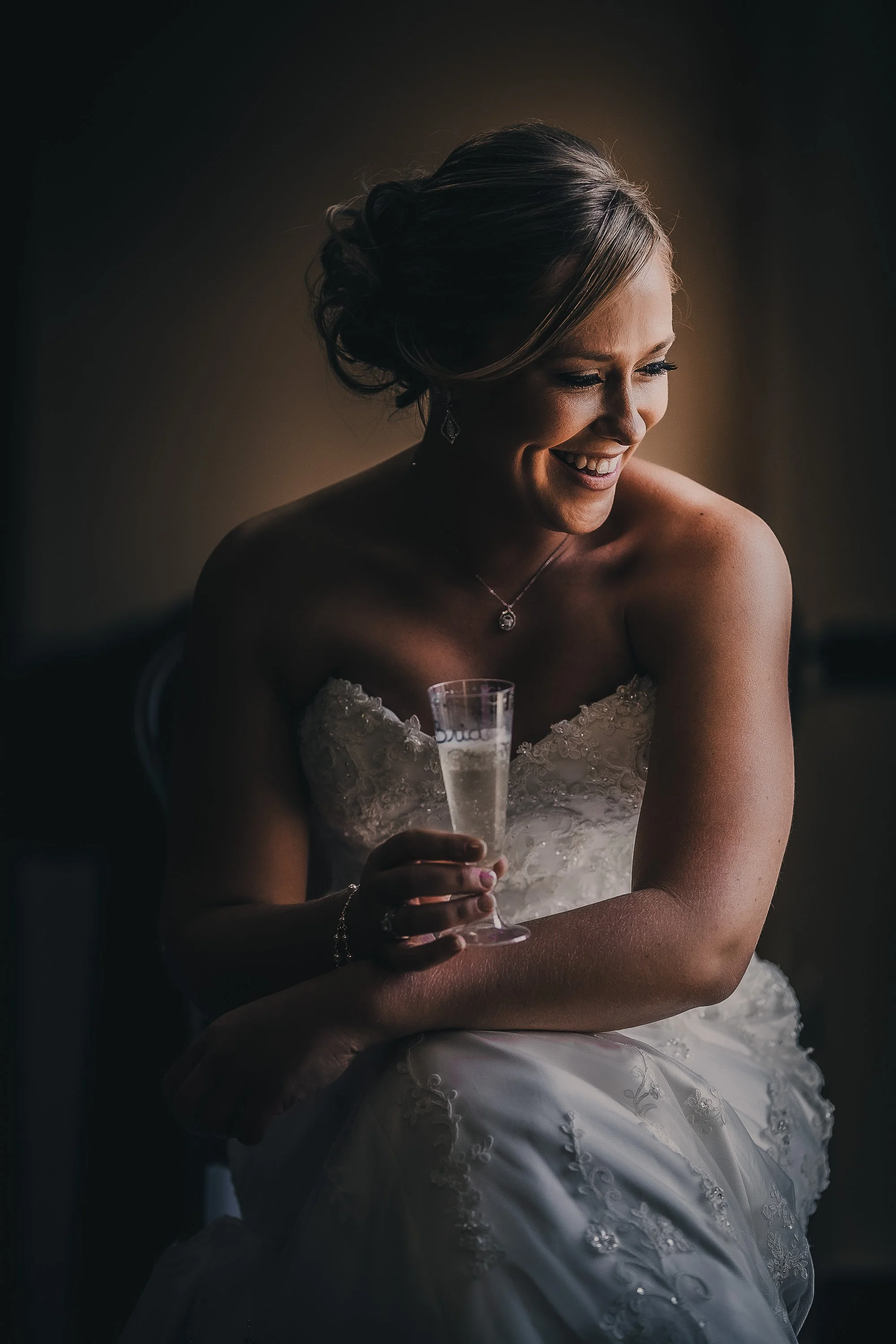 A bride in a strapless wedding gown, smiling while holding a glass of champagne, sitting indoors.