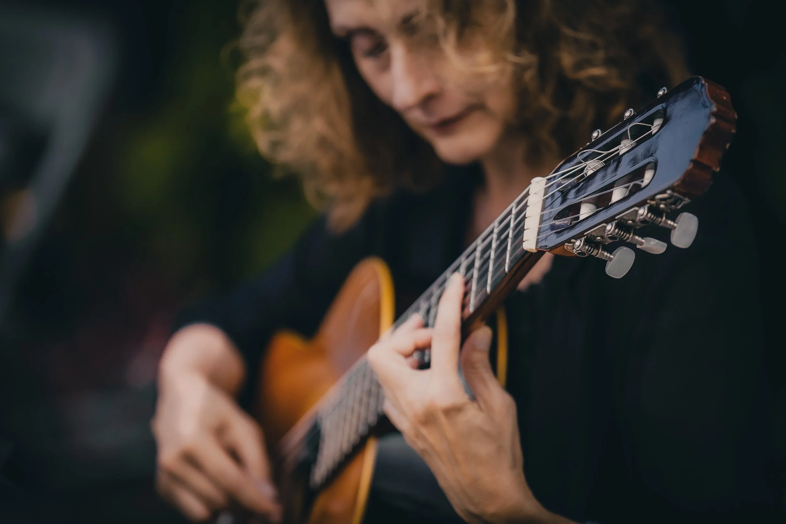 Close-up of a woman playing an acoustic guitar outdoors. She has curly hair and is wearing dark clothing, with a blurred background.