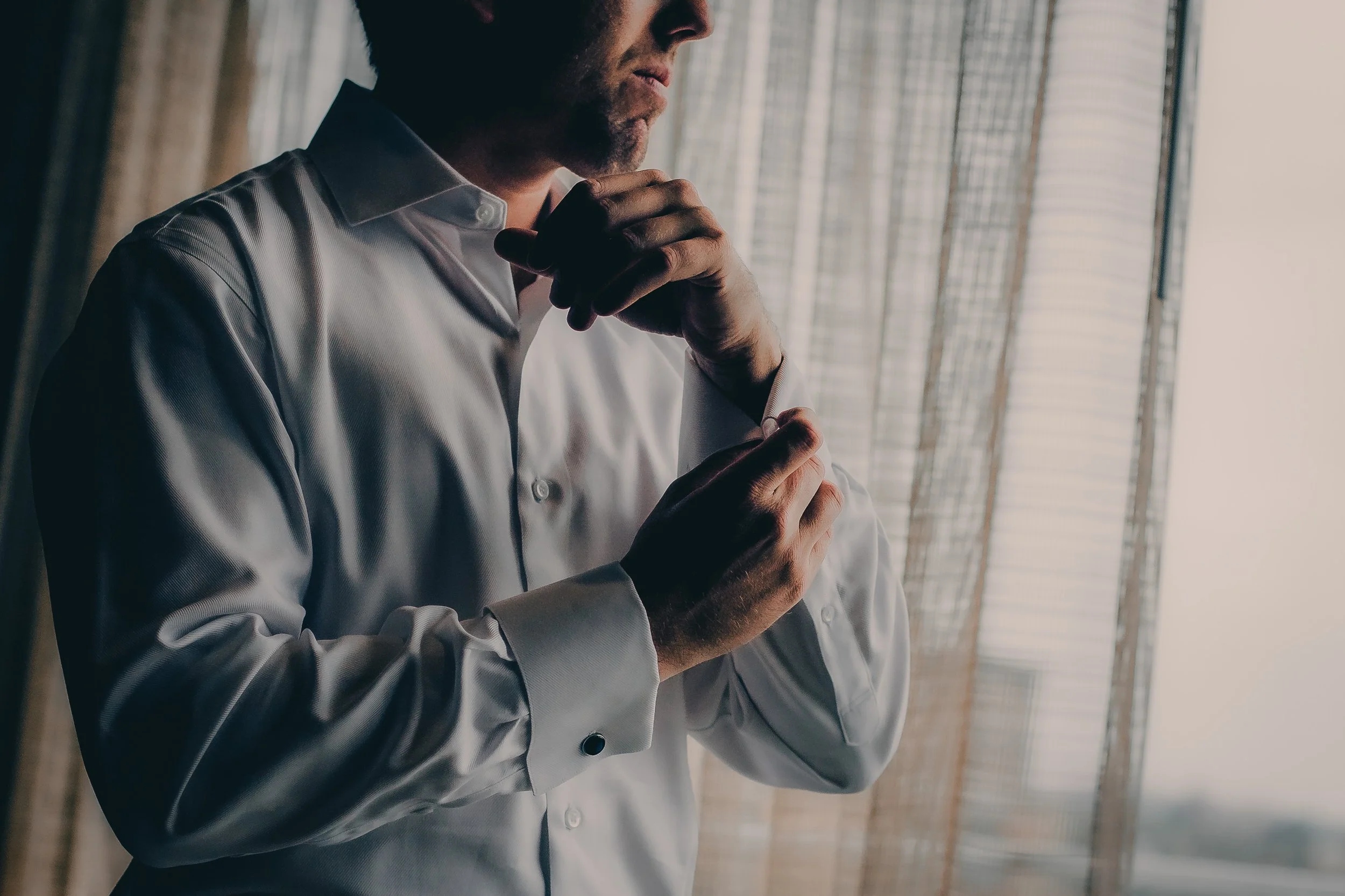 A man is adjusting his cuff links while standing near a window with blinds, dressed in a white dress shirt.