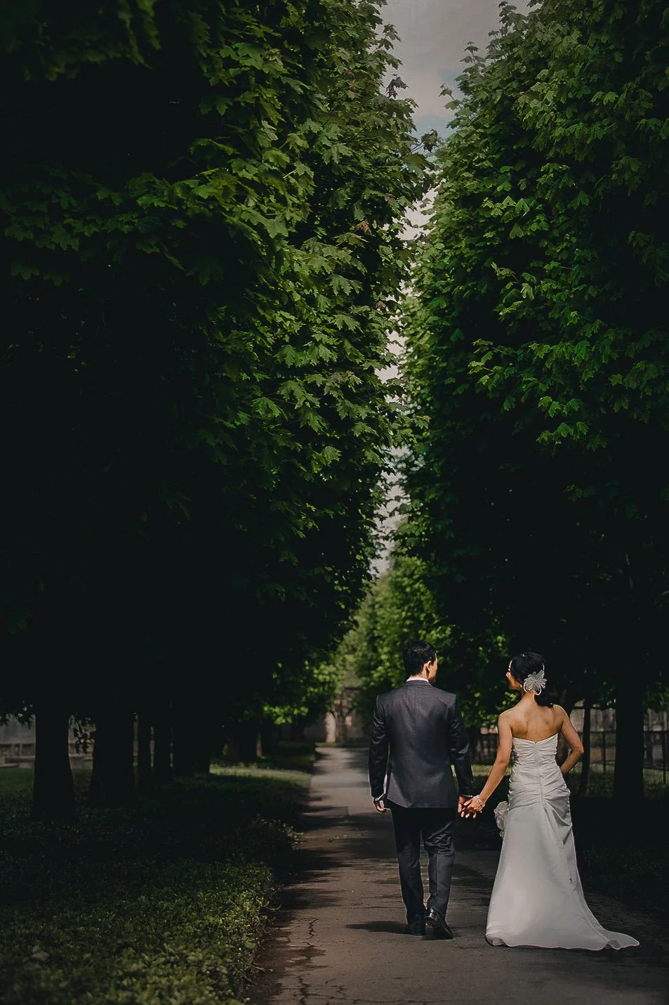 A newlywed couple holding hands and walking down a tree-lined path, with lush green trees on either side under a cloudy sky.