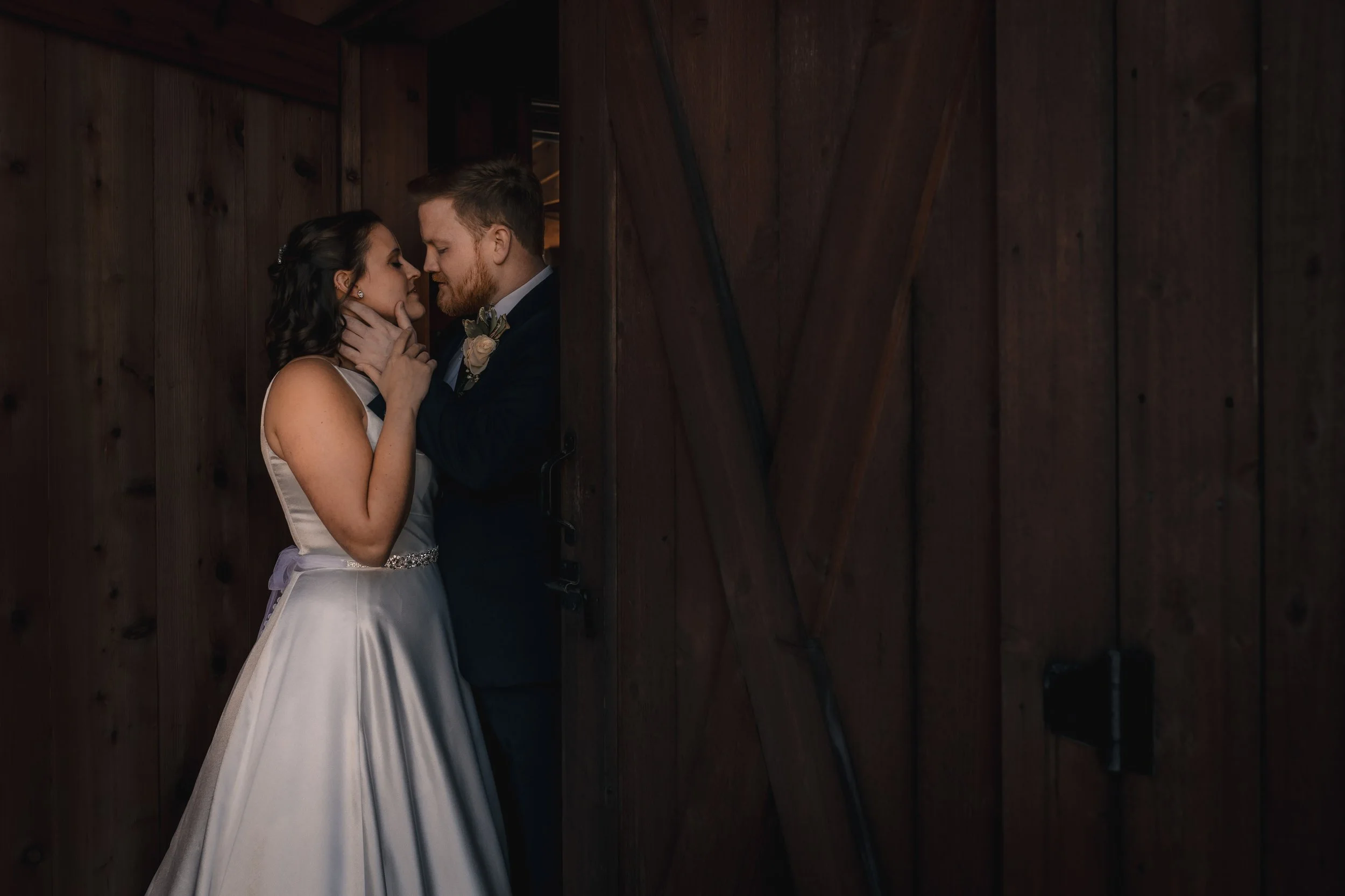 A bride and groom share a romantic moment in a wooden barn, with their faces close, touching noses, and the bride gently holding the groom's chin.